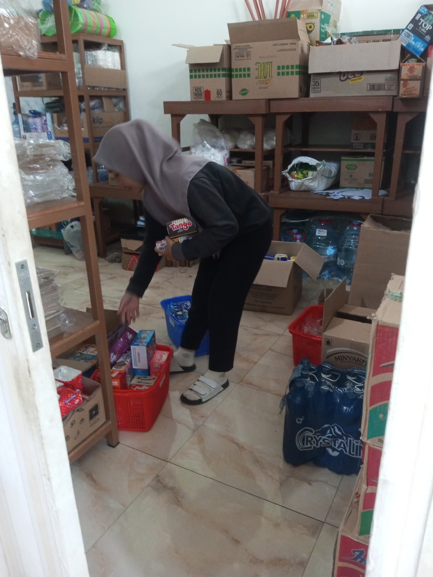 The image depicts an indoor scene, likely a storage area or a small shop, filled with various goods. A person wearing a grey hijab and dark clothing is visible, reaching into a red basket. The person is holding a box of snacks, possibly cookies, with a label that appears to say "Tango". The basket contains several other packaged food items.
The environment is characterized by wooden shelves stacked with cardboard boxes and plastic-wrapped items. Several cases of bottled water are also present on the floor. The flooring is tiled. The overall impression is one of a functional space where goods are stored or organized. The presence of food items and storage shelves suggests a commercial or domestic storage context. There are no clear indicators of an outdoor setting or a specific city like Andongsili, Indonesia, within the visual information provided.