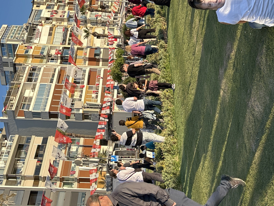 The image captures a street scene in İzmir, Türkiye, specifically at the coordinates 38.438 latitude, 27.141 longitude. It shows a group of people standing on a grassy area, likely a park or a promenade, adjacent to a street lined with multi-story apartment buildings. The buildings are adorned with numerous flags, primarily red with white lettering, and some white and red flags, possibly political or celebratory in nature. The people, diverse in age and attire, appear to be engaged in conversation, perhaps observing something beyond the frame. There are also a few people seated on the grass. The overall scene depicts a typical day in the city, with people going about their daily lives. The buildings, flags, and greenery suggest a vibrant and bustling urban setting.