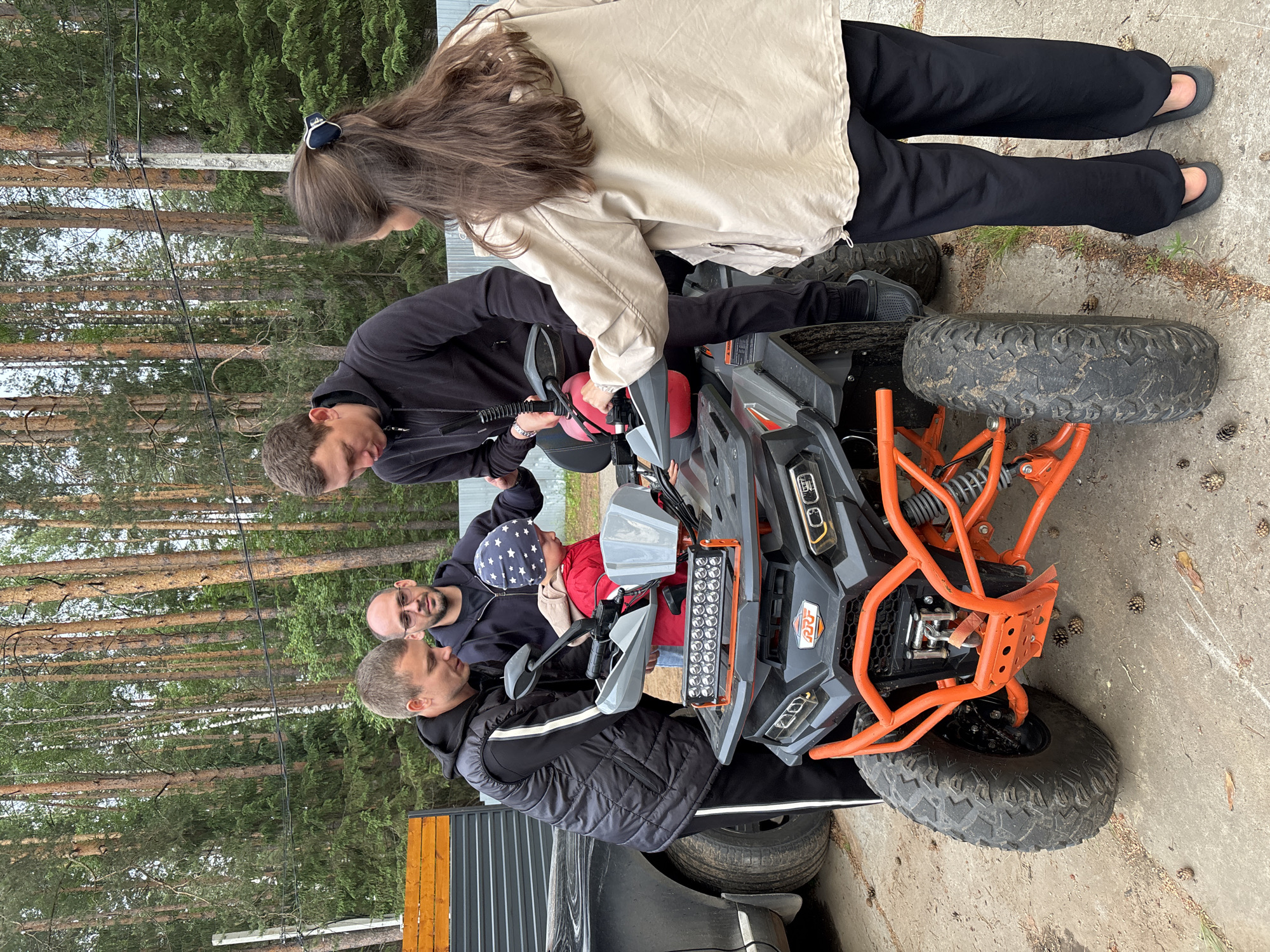 The image displays four individuals—three adults and one child—gathered around an all-terrain vehicle (ATV) on a paved surface, situated at the edge of a coniferous forest, consistent with landscapes found in areas like Chaykovsky, Russia. **People:** * A male individual on the far left, with short brown hair, wears a black puffer vest over a black long-sleeved shirt, black athletic pants with white stripes, and dark closed-toe footwear. He is looking down at the ATV's front section. * To his right, a second adult male with a bald head, dark beard, and eyeglasses wears a dark long-sleeved shirt and blue jeans. He is observing the first male. * A young child, appearing to be a toddler or preschooler, is positioned centrally between two adult males, wearing a red jacket and a blue knit hat with white stars. The child's hands are near the ATV's handlebars. * A third adult male, to the right of the child, has short brown hair and wears a black hooded sweatshirt, black pants, and black Crocs. He is looking down at the ATV near the handlebars. * On the far right, an adult female with long brown hair, secured with a dark hair clip, wears a light tan/beige long-sleeved jacket, black wide-leg pants, and dark flip-flops. She is standing beside the ATV, her right hand resting on its rear seating area. **Subjects & Objects:** * The central object is a dark grey and black ATV with prominent orange frame components, including the front bumper and suspension arms. It is equipped with large, knobby off-road tires, a multi-LED light bar mounted on the front rack, and the brand "RRF" visible on its front. A red plastic fuel canister is visible near the handlebars, and a winch is integrated into the front bumper. * A dark-colored automobile (partially visible) is parked behind the first male on the left. * The ground is paved concrete, scattered with numerous pinecones and some dry organic debris. * In the background, a dense forest of tall, slender coniferous trees (primarily pines) is visible. A concrete utility pole with electrical lines runs vertically through the scene in the background. **Actions & Interactions:** The adults are gathered around the ATV, appearing to be observing or preparing the vehicle. The child is positioned in the center, seemingly engaged with or being shown the ATV, with the adult males leaning over the front section. The