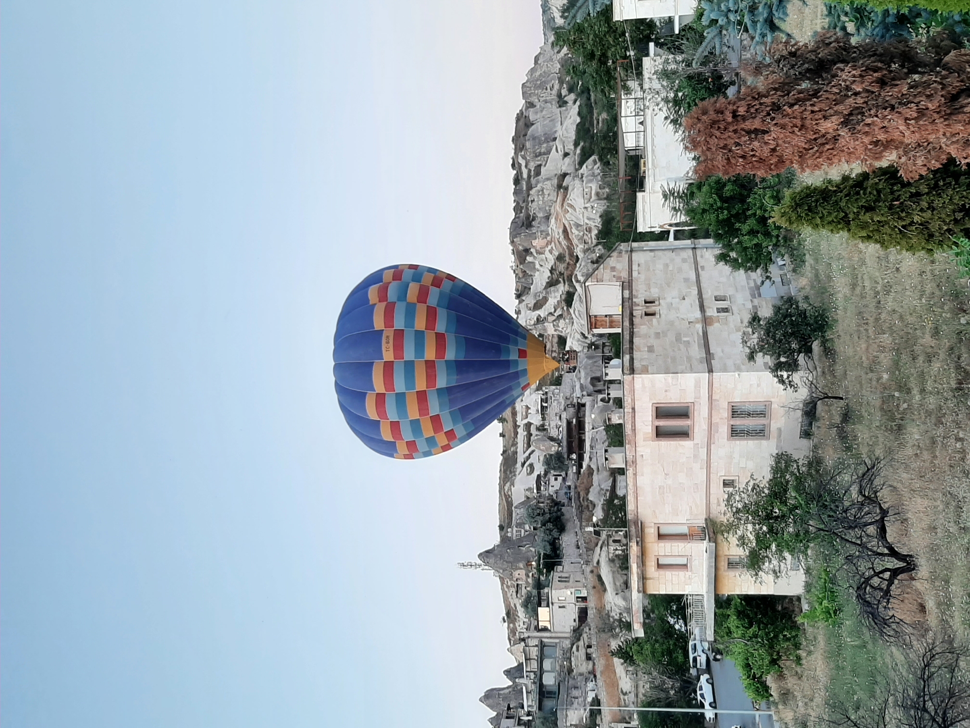 A hot air balloon, featuring blue, yellow, red, and orange horizontal stripes, is airborne over the town of Göreme, Turkey. The balloon is positioned slightly to the left of the frame's vertical center, against a clear, pale blue sky. Below the balloon, traditional Göreme architecture is visible, characterized by stone buildings with flat roofs and numerous windows, some featuring metal grates. Some structures appear to be carved into the soft rock formations that are characteristic of the region. A few trees, one with dry, leafless branches and others with green foliage, are scattered throughout the foreground and midground. The terrain is sloped and appears to be covered with dry grass and scattered wildflowers. In the distance, more rock formations and buildings are visible, along with what appears to be a telecommunications tower atop a rocky outcrop. A road with a few parked cars is also discernible to the left of the frame. The overall scene depicts a typical morning in Göreme, known for its hot air balloon rides over its unique geological landscape.