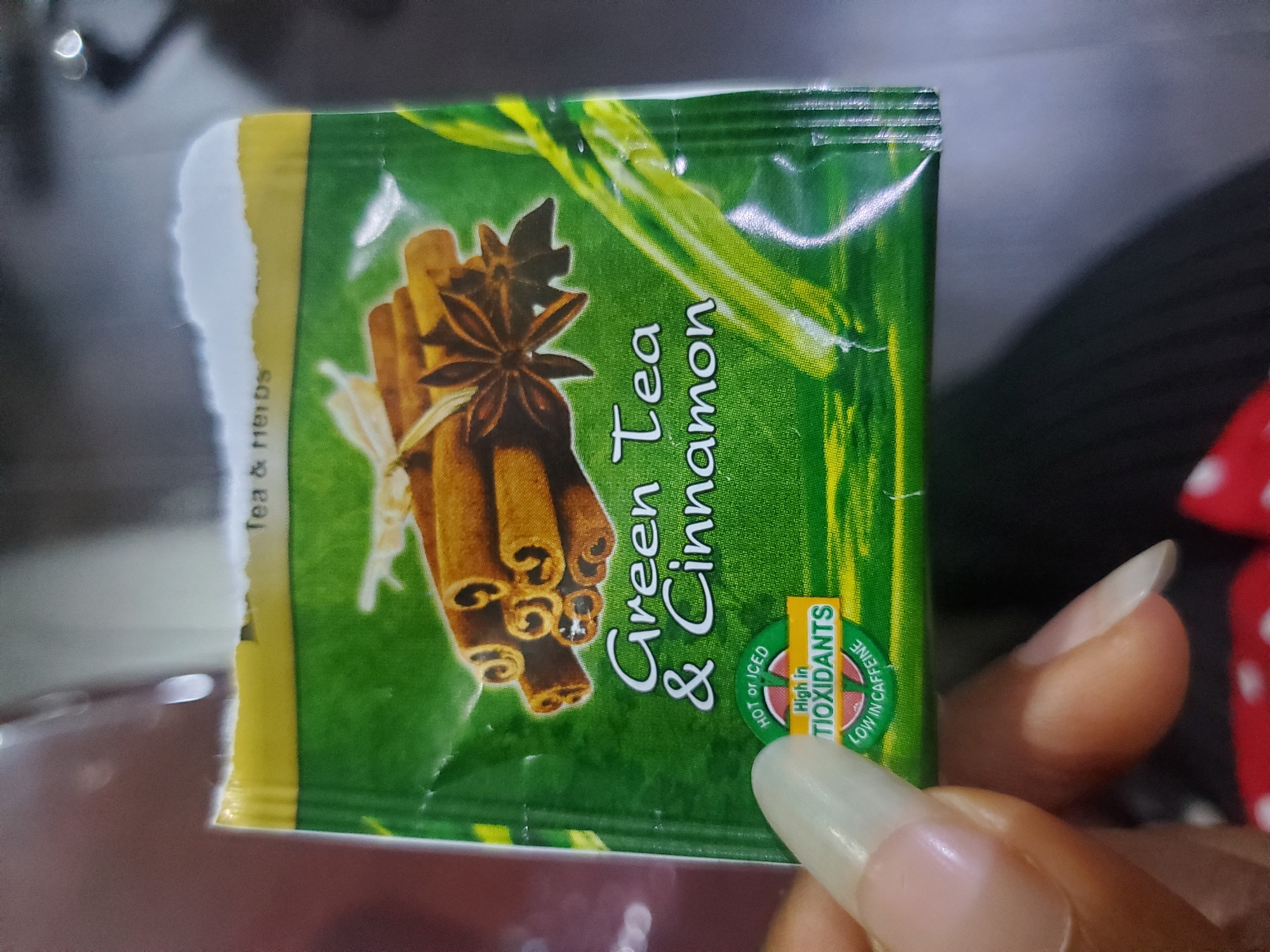 A hand with long, light-colored fingernails is holding a green tea bag packet. The packet has an image of cinnamon sticks and star anise on it, and text that reads "Green Tea & Cinnamon." There is also text indicating it is "High in TIOXIDANTS" and "LOW IN CAFFEINE." The background is out of focus, but appears to be an indoor setting. The image is not specific to Gwagwada, Nigeria.
