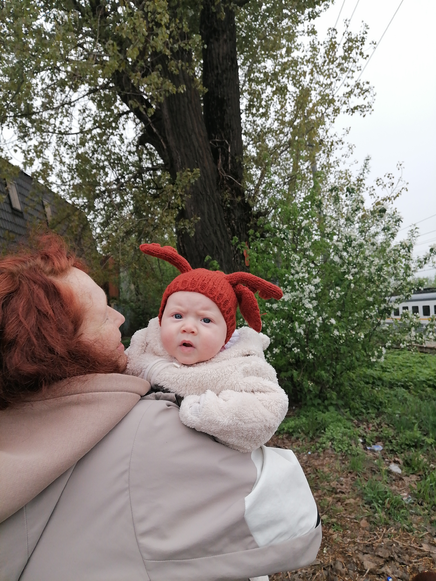 A woman with red hair holds a baby in a white, fuzzy jacket and a red knit hat with antennae-like protrusions. The baby is looking directly at the camera. Behind them, there is a tree with bare branches and a bush with white flowers. Further back, a train is visible. The location is Юдино, Russia, as evidenced by the train type and general landscape which are common in the region. The environment appears to be a slightly unkempt outdoor space.