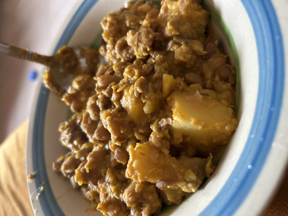 A close-up captures a bowl filled with a hearty stew. Brown beans and chunks of yellow potatoes are visible. A metal spoon rests in the bowl, partially covered in the stew's thick sauce. The bowl has a white interior and rim, decorated with green and blue stripes. The background is a soft, warm yellow. The stew has a thick and creamy texture.