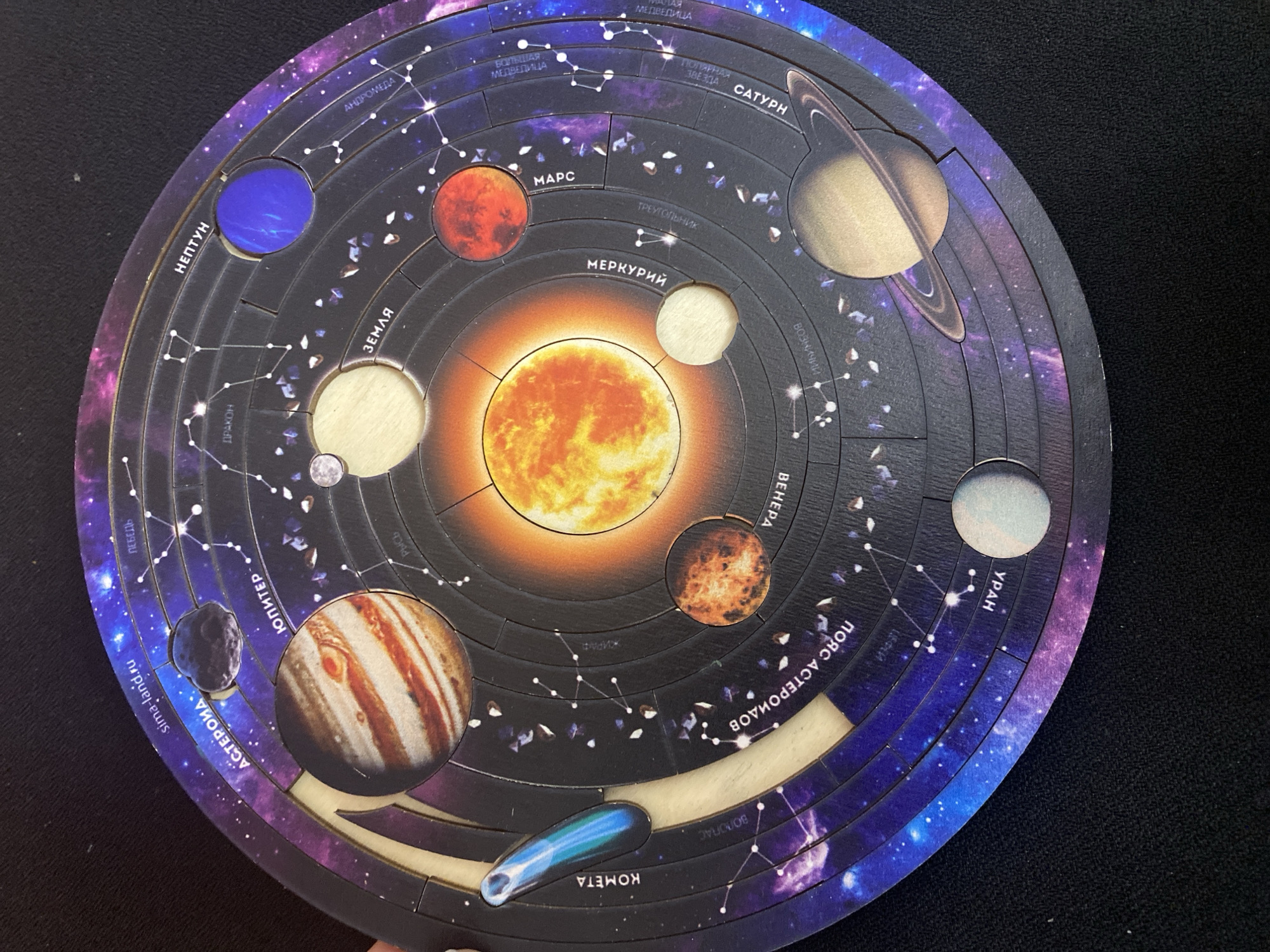 This image captures a circular, wooden educational puzzle representing the solar system, set against a dark background, likely a table or cloth. The central subject is a vibrant orange Sun, around which concentric orbits are carved, depicting the paths of planets and other celestial bodies. Many of the planet and body pieces are currently removed, showing light-colored, cut-out sections beneath, while others are in place.
The puzzle features detailed illustrations of the Sun, planets, an asteroid, and a comet. The outer edges of the puzzle are adorned with a depiction of a starry night sky, complete with constellations marked by lines connecting stars.
Visible text, primarily in Russian, labels various elements:
* **Planets:** МЕРКУРИЙ (Mercury), ВЕНЕРА (Venus), ЗЕМЛЯ (Earth), МАРС (Mars), ЮПИТЕР (Jupiter), САТУРН (Saturn), УРАН (Uranus), and НЕПТУН (Neptune).
* **Other celestial bodies:** ПОЯС АСТЕРОИДОВ (Asteroid Belt), АСТЕРОИД (Asteroid), and КОМЕТА (Comet).
* **Constellations/Stars:** Several constellations are named, including АНДРОМЕДА (Andromeda), БОЛЬШАЯ МЕДВЕДИЦА (Ursa Major), ПОЛЯРНАЯ ЗВЕЗДА (Polaris), ТРЕУГОЛЬНИК (Triangulum), ВОЗНИЧИЙ (Auriga), ДРАКОН (Draco), РЫСЬ (Lynx), ЖИРАФ (Camelopardalis), ЛЕБЕДЬ (Cygnus), and ВОЛОПАС (Boötes).
* A website address, "sima-land.ru," is also visible on one of the outer pieces.
The puzzle pieces are distinct cutouts, suggesting an interactive toy designed for learning about astronomy. The vivid colors and detailed illustrations make it an engaging educational tool. No specific time of day or location cues are apparent other than the object itself. The Russian text aligns with the given context of Saint Petersburg, Russia, suggesting a product from that region.