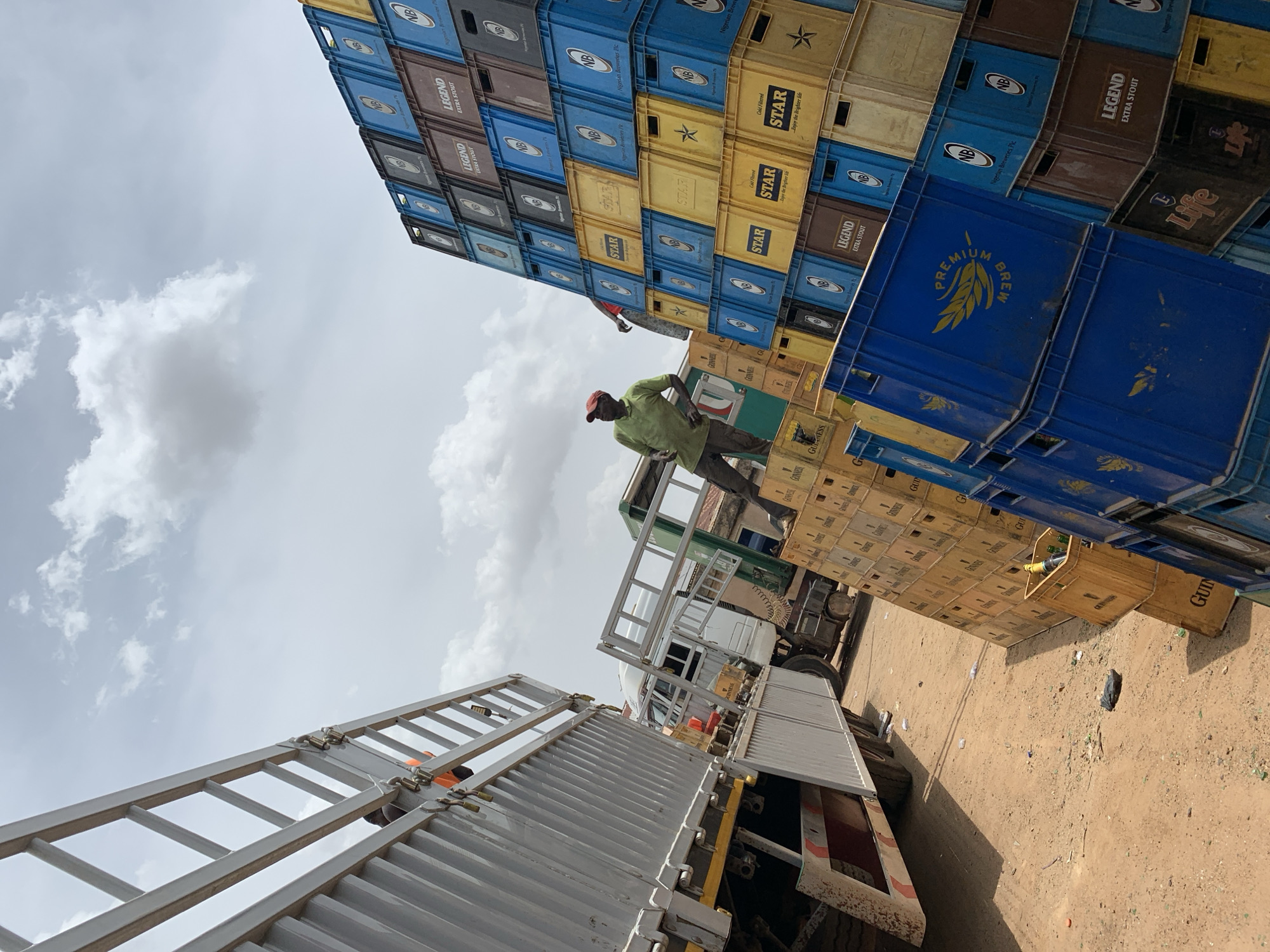 The image depicts a man in Bukuru, Nigeria, climbing a stack of beer crates next to a truck. The crates are stacked high, composed of brands like Guinness, Star, and Legend. The environment appears to be an open lot with a sandy ground. A ladder is attached to the side of the truck. The sky is overcast with clouds.