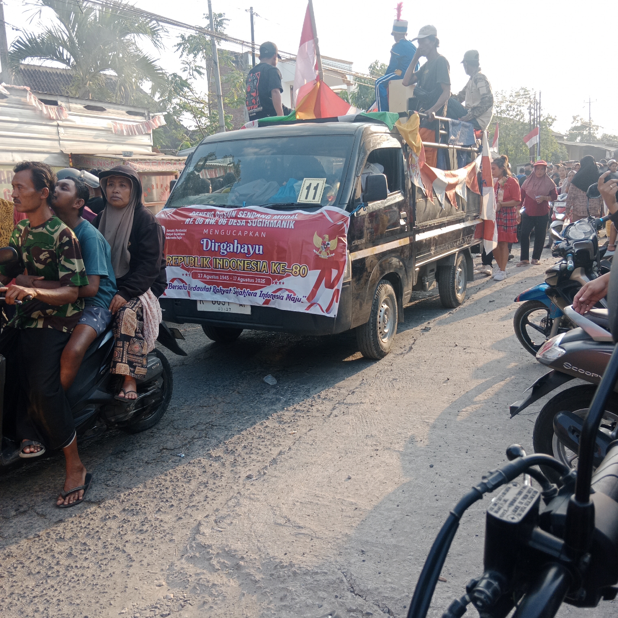 The image depicts a street scene in Tegalrejo, Indonesia, likely during a procession or celebration. A black pickup truck, adorned with red and white banners, is the central focus. The banner reads "Dirgahayu Republik Indonesia Ke-80," indicating a celebration of Indonesia's 80th Independence Day. The truck carries several people, some in what appear to be marching band uniforms with hats and blue jackets, while others are dressed casually.
In the foreground, two men are seated on a motorcycle. The man in front wears a camouflage shirt and a black head covering, while the person behind him wears a blue t-shirt. To the right of the truck, a woman in a brown hijab and a patterned skirt is visible, alongside other individuals and a blue motorcycle. The street is paved with asphalt and concrete, and the scene is bathed in the warm light of what appears to be late afternoon. Palm trees and buildings line the background, suggesting a residential or semi-urban environment. The overall atmosphere suggests a community event or parade.