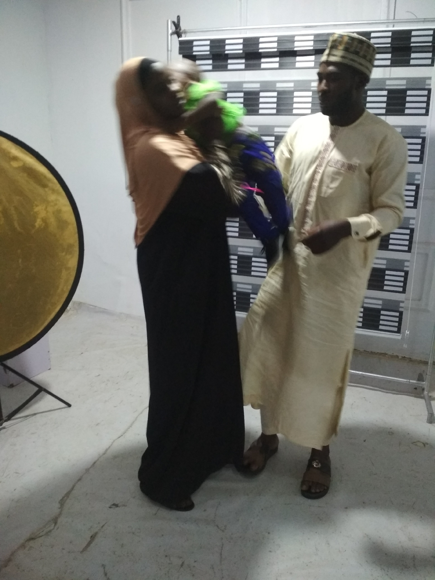 A photograph shows a woman holding a baby, standing next to a man. The woman is wearing a black robe with a brown headscarf. The baby is wearing green and blue clothes. The man is wearing a light-colored long robe with a patterned cap, and sandals. In the background is a wall with a black and white patterned divider and a gold reflector. The location is Unknown, Unknown.
