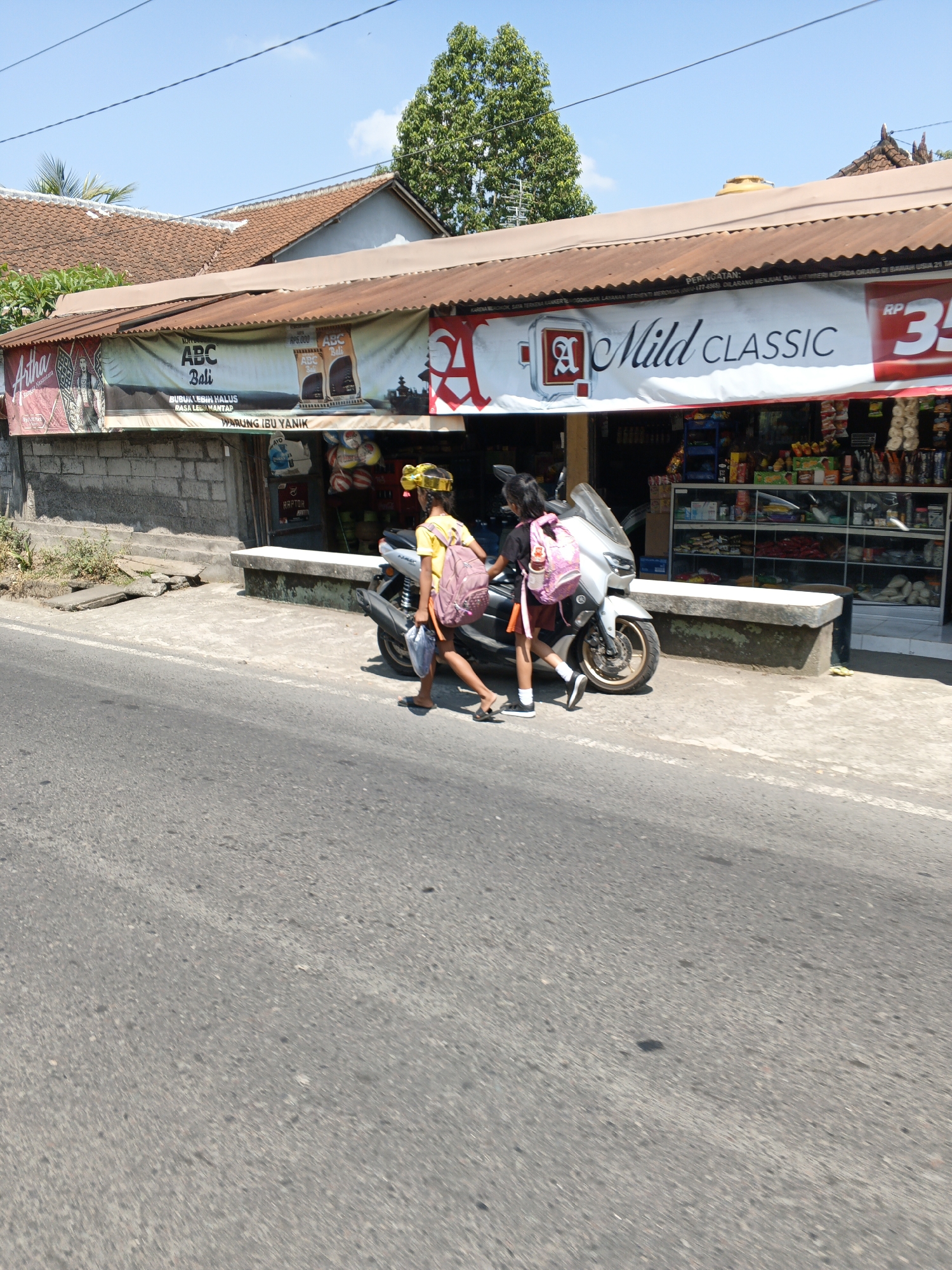In Ubud District, Indonesia, two young girls are walking past a small roadside store. The girl on the left, wearing a yellow shirt and a yellow hair accessory, carries a pink backpack. The girl on the right, wearing a dark t-shirt and a reddish-brown skirt, also has a pink backpack. They are walking alongside a motorcycle. The store, with a corrugated metal roof, displays banners advertising various products, including "ABC Bali" and "Mild CLASSIC." The interior of the store is visible through glass display cases filled with merchandise. The scene is set against a backdrop of a clear blue sky and lush green trees.