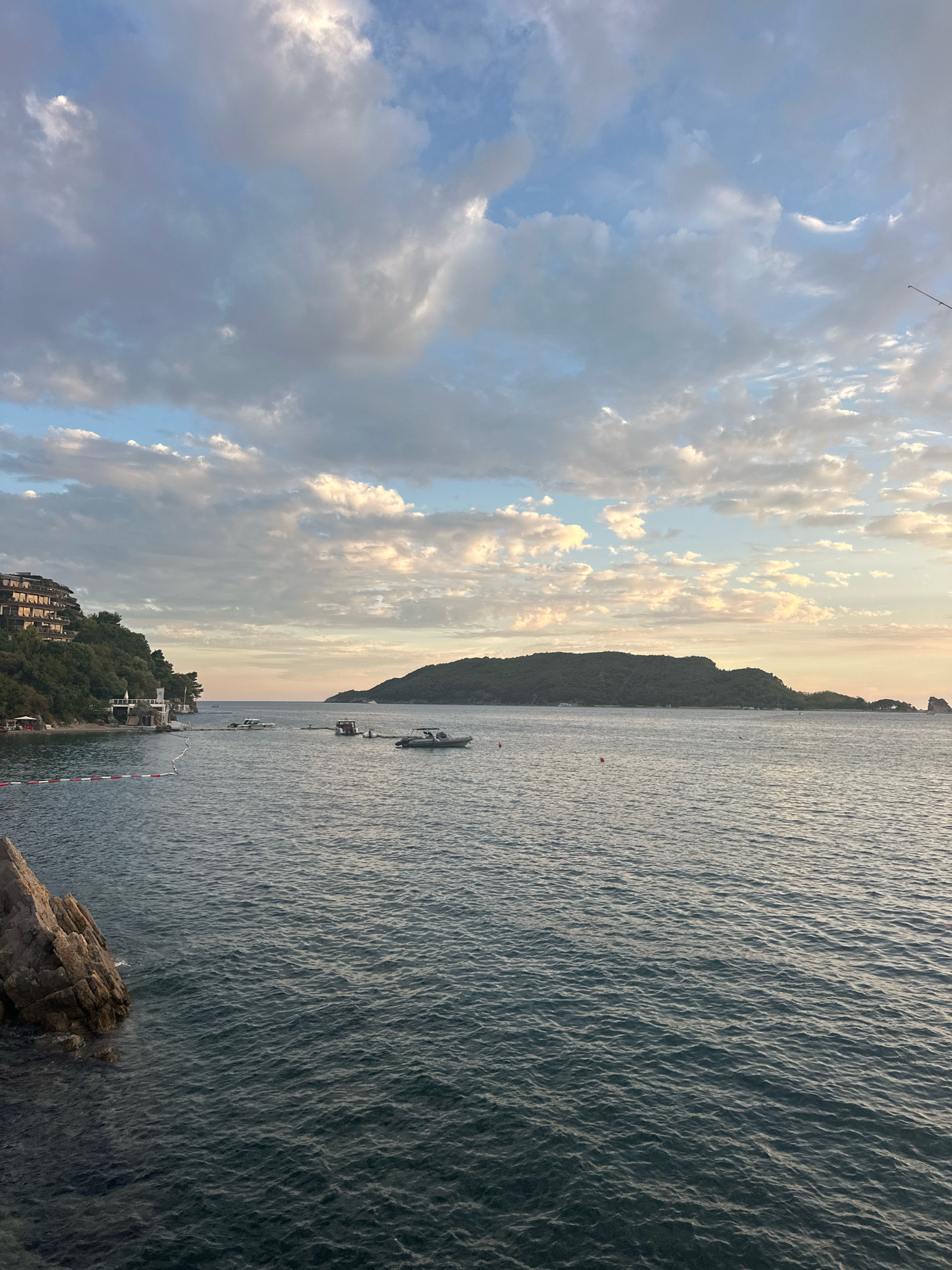 The image depicts a scenic view of the Adriatic Sea at dusk, likely near Budva, Montenegro. In the foreground, dark blue water is visible with gentle ripples. To the left, a rocky outcrop juts into the sea. Further back, a beach area is demarcated by a red and white buoy line. On the left side of the frame, a multi-story building with curved architecture is nestled into a green, tree-covered hillside. Several boats are scattered across the water, including a white motorboat closer to the shore and a larger vessel further out. In the distance, a large, verdant island dominates the horizon, with smaller rocky formations visible to its right. The sky is a mix of pale blue and soft grey clouds, with hints of golden light suggesting either sunrise or sunset. The overall atmosphere is tranquil and picturesque.