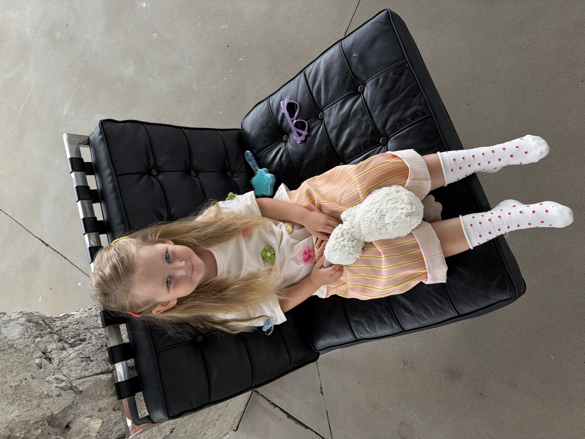 A young girl with long blonde hair lies on a black leather Barcelona chair. She is wearing a white t-shirt with flower embellishments, a pink and yellow striped skirt, and white socks with red polka dots. She is holding a white stuffed animal. Beside her on the chair are purple sunglasses and a blue hairbrush. The chair is situated on a gray concrete floor. There is a gray textured wall behind the chair. There is no indication that this image was taken in Saratov, Russia.