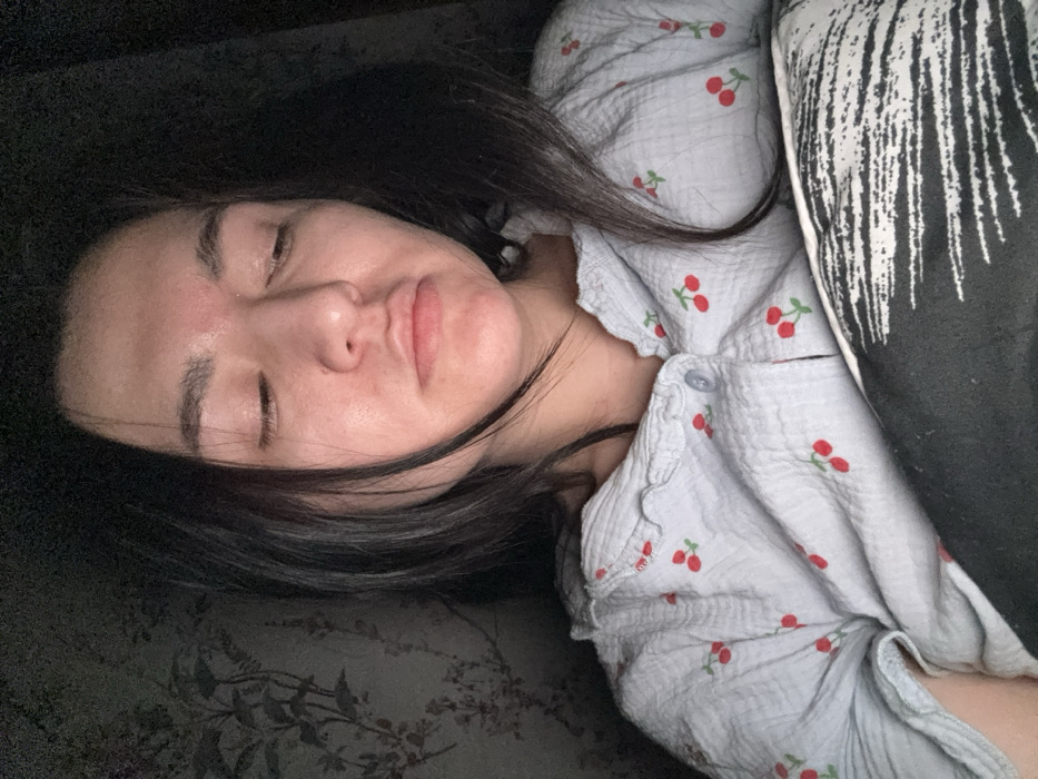The image shows a female individual indoors, likely in Kurovskoye, Russia, resting in a dimly lit setting. Her head is tilted slightly to her right, and her eyes are closed, indicating sleep or repose. Her lips are slightly parted. She has dark hair, which is loose and falls around her face and shoulders. The individual is wearing a light-colored, possibly pale blue or grey, textured button-down shirt with a ruffled collar and a repeated pattern of small red cherries with green stems. A dark pillow or blanket with a white abstract pattern is visible in the lower right portion of the frame, partially covering her torso. The background on the left side reveals a wall adorned with a subtle, dark floral or botanical pattern, consistent with wallpaper. No other individuals or active interactions are visible.