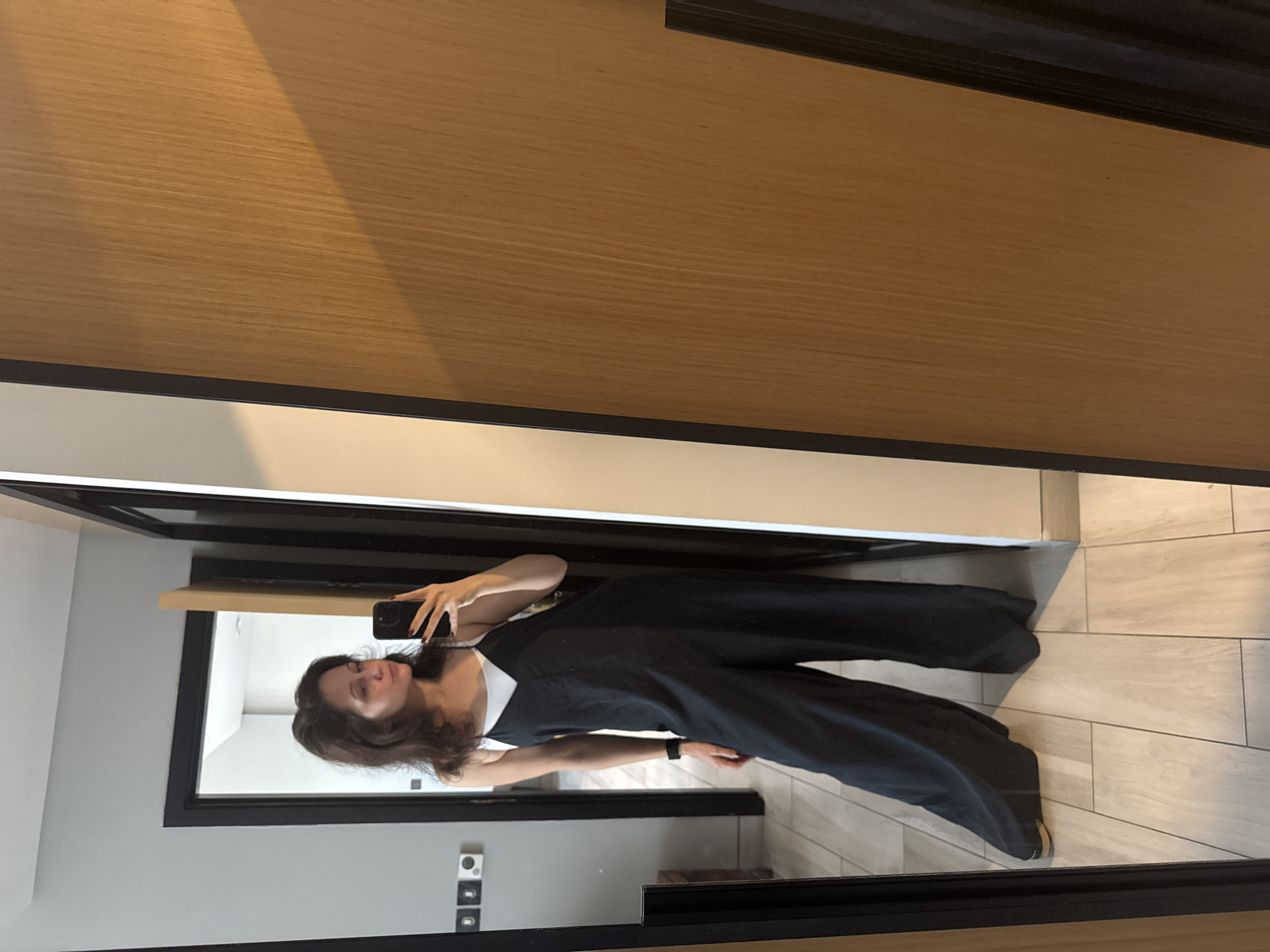 A woman is taking a selfie in a mirror. She is wearing a dark jumpsuit with a white tank top underneath. She is holding a smartphone in her right hand. Her hair is dark and shoulder-length. The background is a room with light-colored walls, light tiled flooring, and wooden panels. A black framed mirror is on the wall, possibly in a hotel room. The location might be in Dubai, United Arab Emirates.
