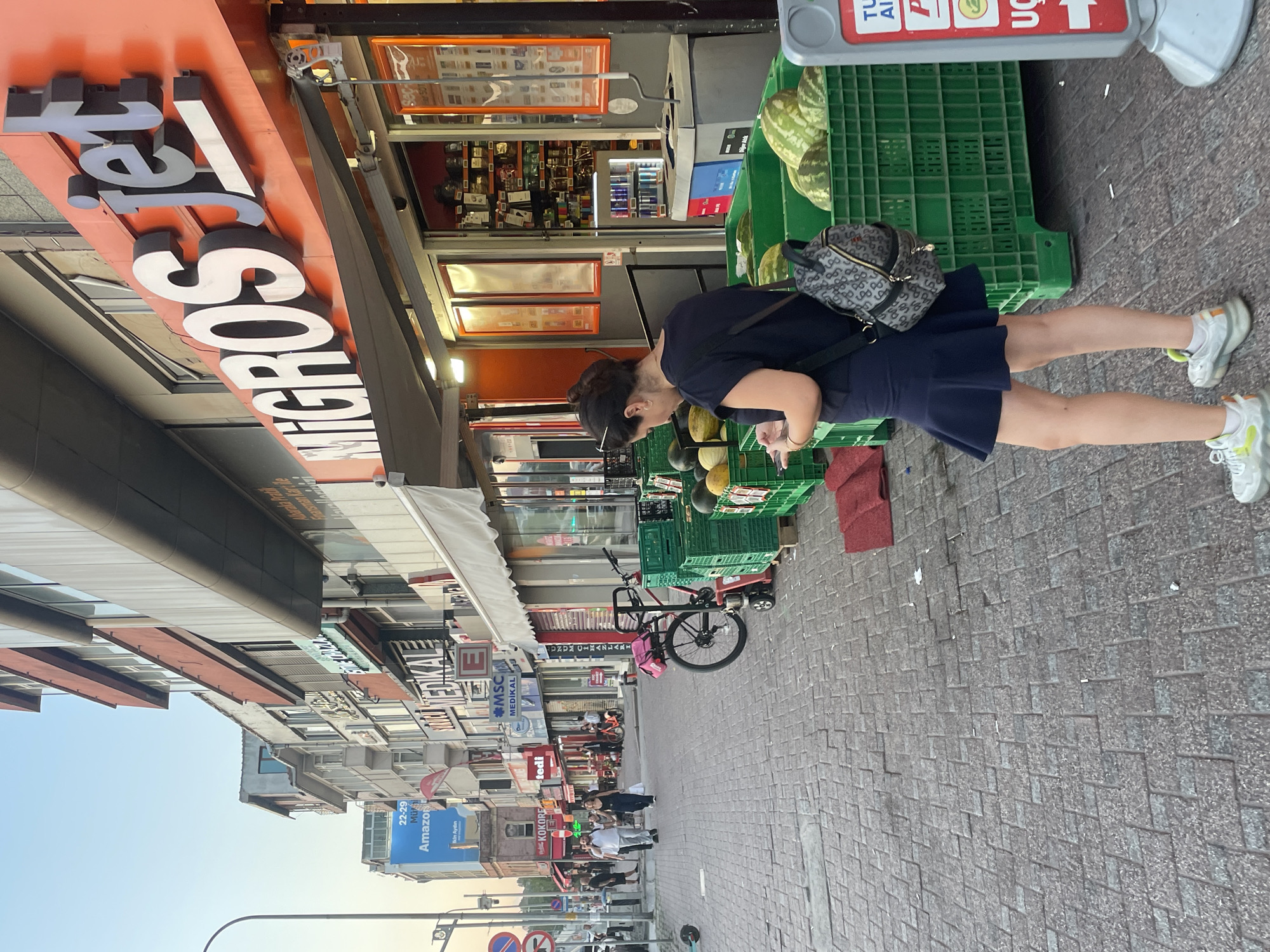 In Kadıköy, Turkey, on a cobblestone sidewalk, a woman stands looking at her phone. She has dark hair pulled back and wears a dark blue skirt and top, with a patterned backpack slung over her shoulder and white sneakers. In front of her, a Migros Jet store is visible with its orange facade and large "Migros jet" signage. Green crates overflowing with watermelons and other fruits are displayed outside the store. Behind the woman, other storefronts line the street, and people are walking in the background. The sky is a clear, pale blue, suggesting late afternoon or early evening.