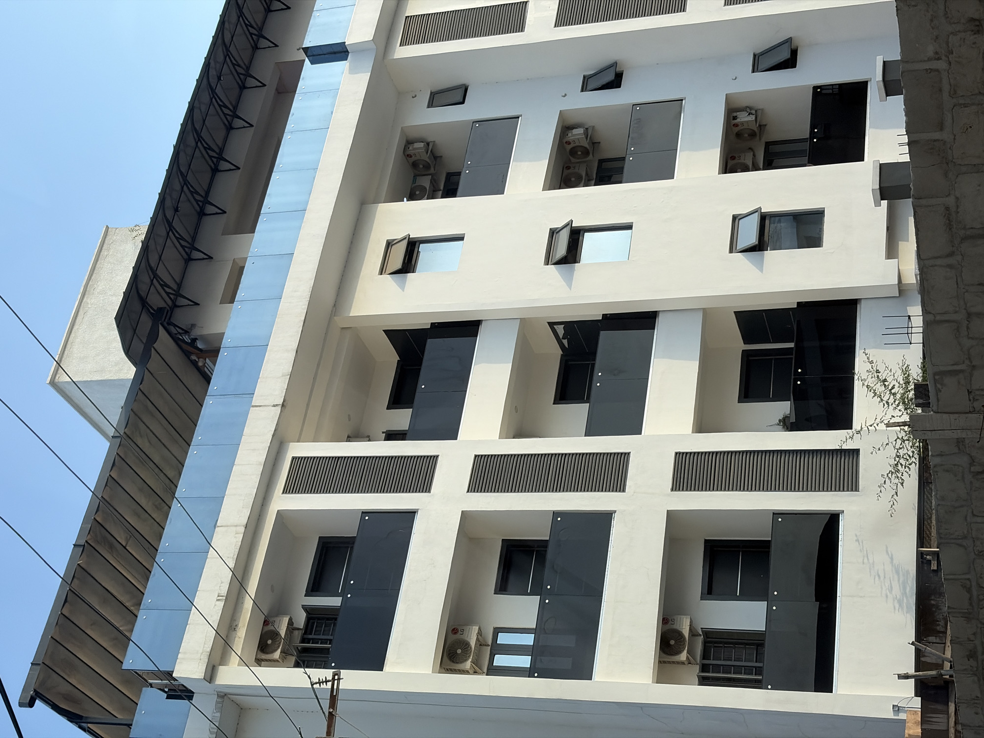 The image captures a low-angle view of a modern, multi-story building. The building is predominantly white with dark windows and rectangular cutouts that give it a distinct, patterned facade. Many windows are partially open. Air conditioning units are visible within some of the window spaces. A glass facade runs vertically along one side of the building. A dark, slatted awning is visible above the glass facade. The sky is pale blue. Power lines are visible in the foreground. The exact location within the city of Moba, Nigeria, is not discernible from the image. There are no visible people.
