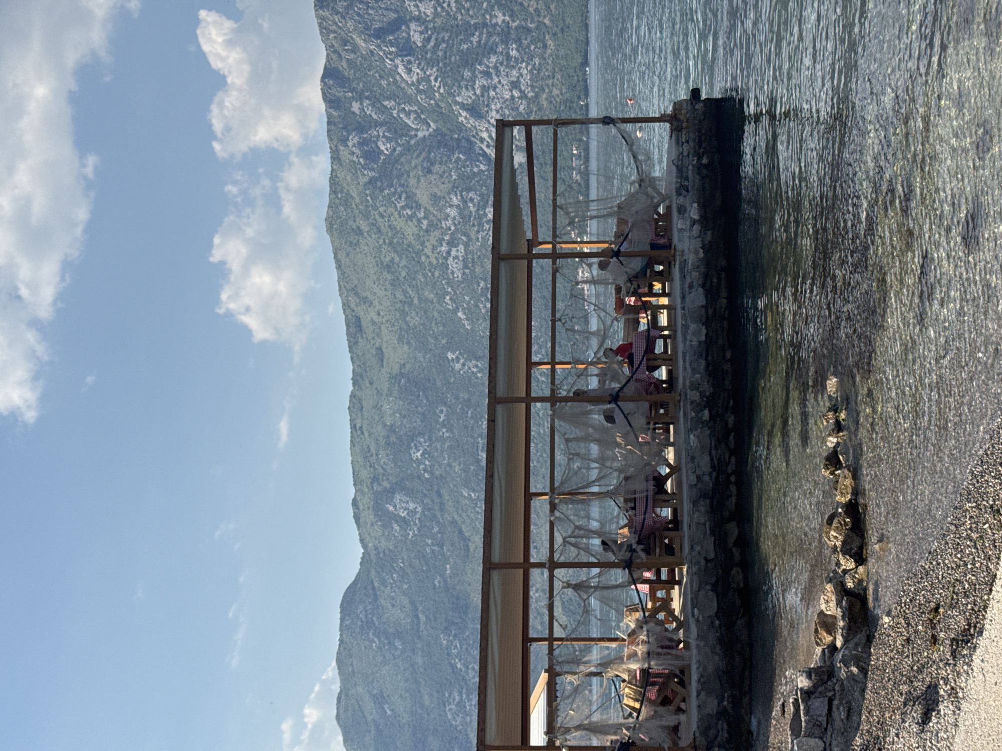 The image captures a waterside restaurant in Prčanj, Montenegro. The restaurant is situated on a stone embankment extending into the bay. Its structure consists of a simple wooden frame supporting a roof, with seating arranged inside. Nets or light fabric hang around the edges. To the left, a mountain rises, partially obscured by a sky with scattered clouds. The water laps at the embankment's edge. Rocks and gravel form the shoreline. The image is vertically oriented.