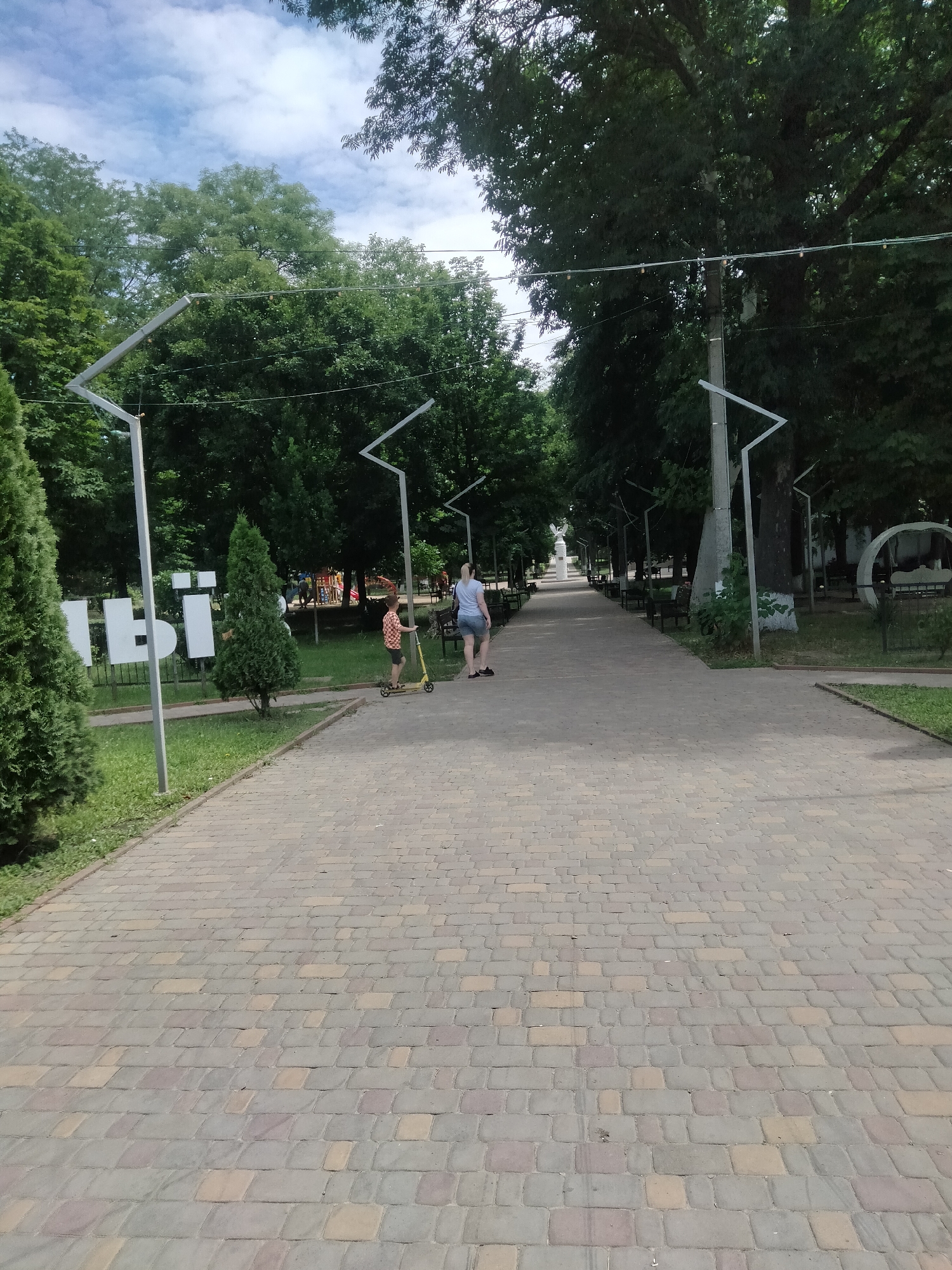 The image shows a park pathway in Ananiv, Ukraine. The pathway is paved with interlocking bricks in a herringbone pattern. On the left side of the pathway, there are large white letters, the first two of which appear to spell "ІВ", with a partial third letter visible. Several large trees with green foliage line the pathway on both sides, and modern-looking, angular lamp posts are also visible. In the mid-ground, two people are walking. A young boy on a yellow scooter is in front and to the left of an adult woman. The woman is wearing a light blue t-shirt and denim shorts. The boy is wearing a red and white checkered shirt and dark shorts. In the background, there are more trees, park benches, and what appears to be a children's playground with colorful equipment. The sky is cloudy with patches of blue.