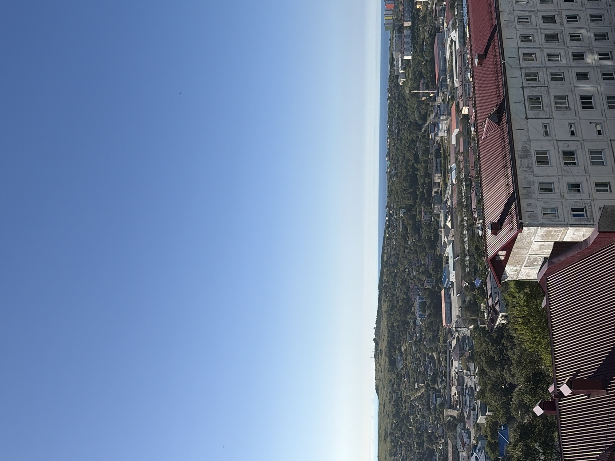 This is a high-angle shot of Korsakov, Russia, on a clear, sunny day. The sky is a vibrant, cloudless blue. In the foreground, a multi-story building with a red, corrugated metal roof and many windows is prominently featured. Adjacent to it, another building with a similar red roof is partially visible.
The middle ground reveals a densely populated urban landscape with numerous buildings, roads, and trees. The buildings vary in size and style, with some appearing industrial and others residential. Green trees are interspersed throughout the city, suggesting a degree of urbanization and natural integration.
In the background, a gentle hill covered in vegetation overlooks the city. Beyond the hill lies the calm, blue ocean, meeting the horizon under the clear sky. The distant coastline is visible, and a few scattered structures can be seen on the hill. The overall impression is of a coastal city on a pleasant day. No people are visible in the frame.