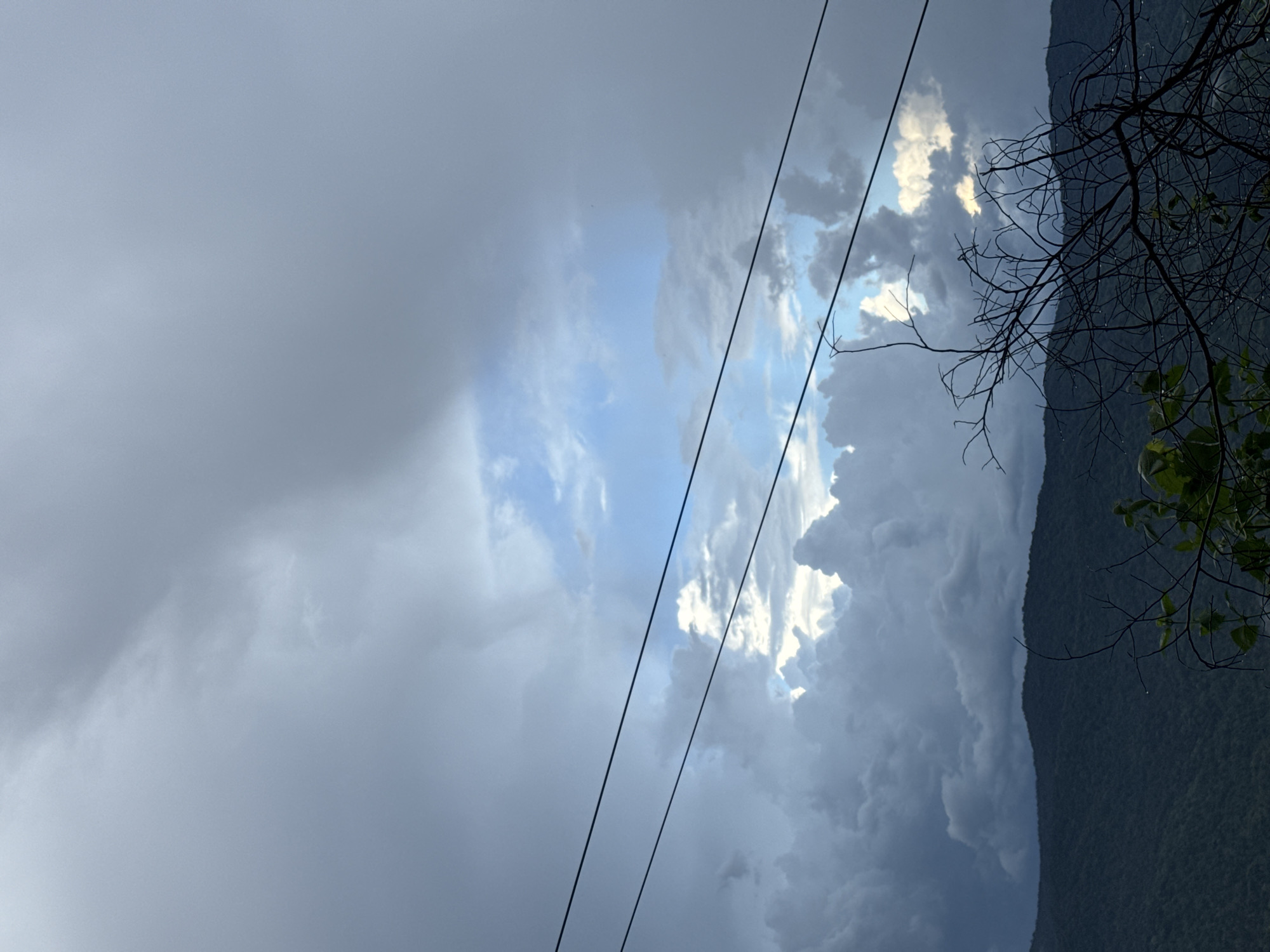 A panoramic view of a vast, overcast sky looms above a dark, mountainous landscape. In the mid-ground, two parallel power lines cut across the frame, their thin, dark forms contrasting with the dramatic cloud formations. The clouds are a mixture of dark, heavy storm clouds and lighter, more diffused masses, with patches of bright blue sky visible through the breaks. The mountains, covered in dense vegetation, form a dark, undulating silhouette against the sky. In the foreground, the bare branches of a tree, with a few sparse leaves, add a touch of delicate detail against the dramatic backdrop of the sky and mountains. The image appears to have been taken in the late afternoon or early evening, judging by the subdued lighting and the ominous cloud cover. The location is specified as Nao Sẻ, Vietnam, suggesting a rural or mountainous region. There are no people or visible actions depicted in the image.