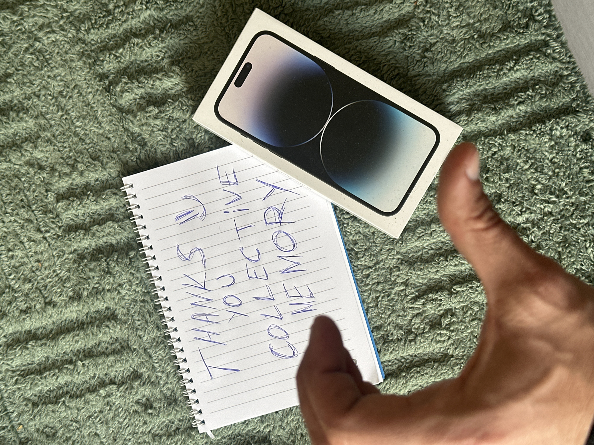 The image presents an indoor scene, likely in Slavyani, Bulgaria, featuring a new iPhone box, a handwritten note, and a person's hand expressing a positive sentiment. The items are arranged on a textured, rumpled green fabric, possibly a blanket or rug.
In the upper right quadrant of the frame, a white iPhone box is prominently displayed. The box shows an image of an iPhone screen featuring a dark blue and purple gradient wallpaper and the distinct "Dynamic Island" notch, indicating a Pro model. Below and to the left of the iPhone box lies a spiral-bound notepad with white lined paper and a blue back cover. Written in blue ink on the notepad are the words: "THANKS YOU COLLECTIVE MEMORY," accompanied by a small symbol that resembles two downward-pointing arrows or a stylized 'U'.
In the lower right, a human hand, appearing to be a right hand, makes a clear "thumbs up" gesture. This gesture, along with the message on the notepad, conveys feelings of satisfaction, gratitude, or approval. The lighting in the image is bright and even, suggesting it was taken during the day.