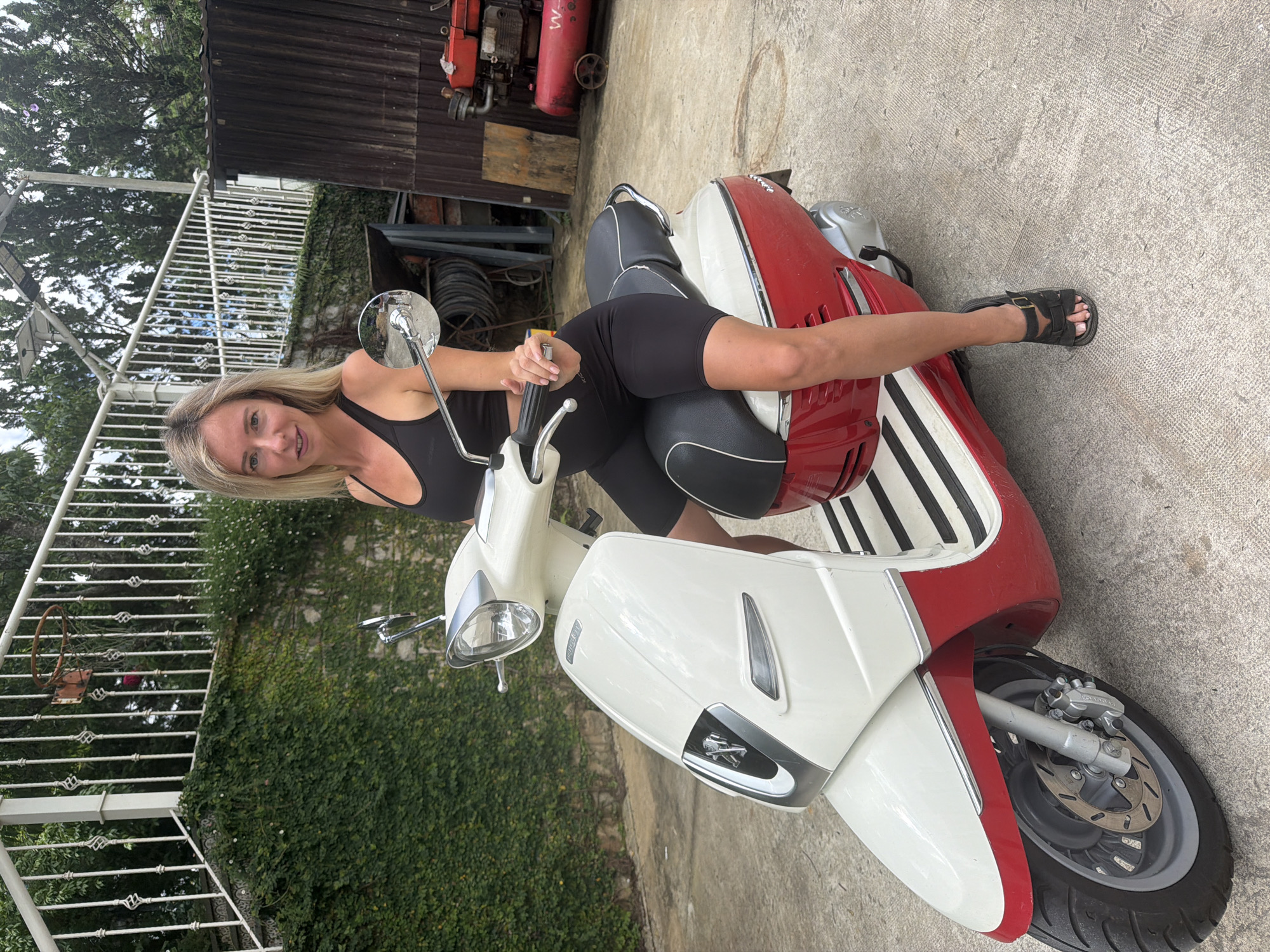 A woman with blonde hair is sitting on a red and white scooter. She is wearing a black tank top and black shorts. She has on black sandals. The scooter is parked on a concrete surface. Behind the scooter, there is a building with a wooden facade and some red equipment. There is also a white metal fence with a vine-covered wall behind it. The scene appears to be outdoors, possibly in a backyard or workshop area. The lighting suggests it is daytime. The woman is smiling and looking at the camera.