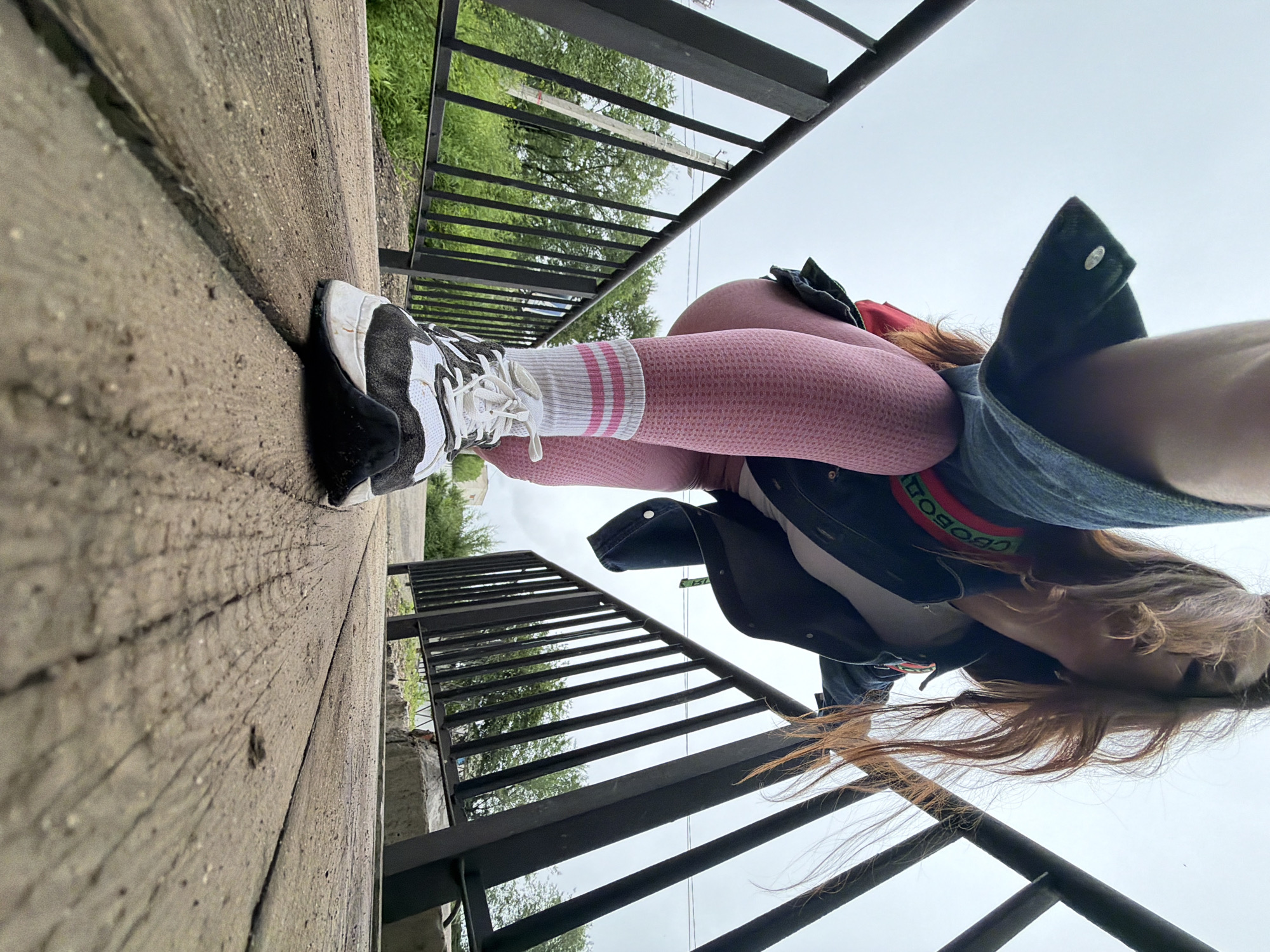 The image depicts a low-angle shot of a young Caucasian female, seen from the knees down, standing on a wooden walkway. She is wearing pink textured leggings, white socks with pink stripes, and black and white athletic shoes. Her denim jacket, with a red and green stripe detail visible on the back, is pulled down, obscuring her torso. Her auburn hair is flowing around her face, which is not fully visible. The walkway appears to be a bridge or elevated path, with black metal railings on either side. Beyond the railings, lush green trees and vegetation are visible, suggesting an outdoor, possibly park-like, setting within the city of Birobidzhan, Russia. The sky is overcast and light gray. The perspective suggests the photo was taken by the subject herself, possibly using a wide-angle lens or a wide-angle setting on her device.
