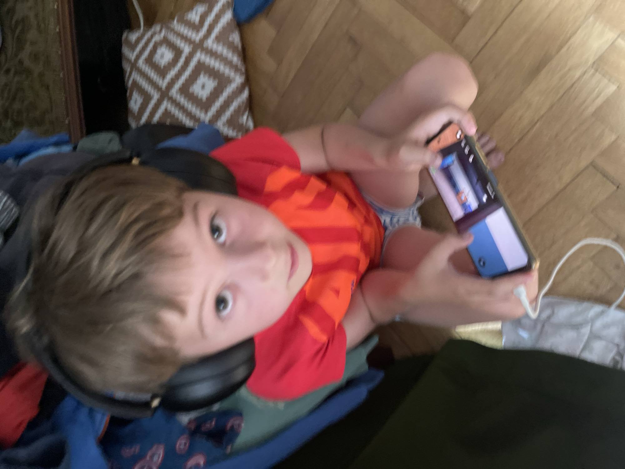 A young boy wearing headphones and a red t-shirt is sitting on a wooden floor, holding a smartphone. The boy is looking at the phone, which appears to be a game or video. The background is cluttered with clothes and a patterned pillow. The lighting suggests it is daytime. The image does not contain enough information to determine the location within the city of Bydgoszcz, Poland.