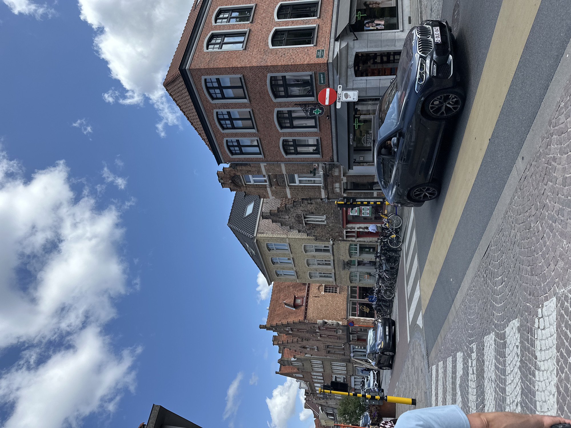 A street scene in Bruges, Belgium, is depicted under a blue sky with scattered white clouds. The foreground shows a cobblestone crosswalk with a yellow stripe marking the pedestrian crossing. A dark grey BMW SUV is driving across the intersection, its license plate reading "2-DLE-847". To the right of the SUV, a red brick building with a dark tiled roof features a sign that says "APOTHEEK" with a green cross emblem. Further down the street, a row of buildings with traditional Flemish architecture lines the left side of the road, displaying various shopfronts and windows. A line of parked bicycles is visible along the sidewalk in front of one of the buildings. A person in a light blue shirt is partially visible on the far left edge of the frame, facing away from the camera. The street is paved with cobblestones and asphalt, and a yellow traffic pole with a light signal stands near the crosswalk.