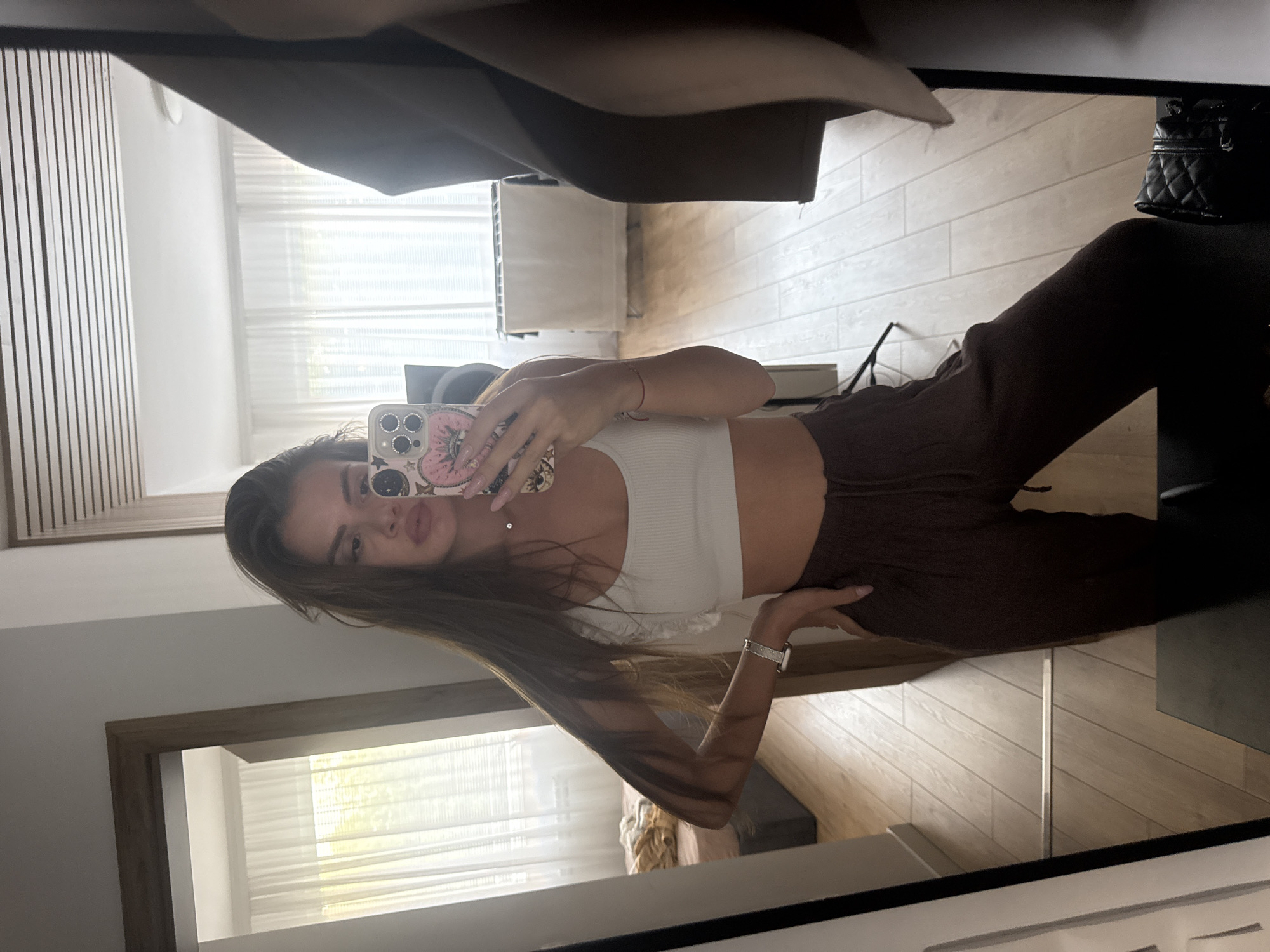 A young adult female is captured in a full-length mirror selfie within what appears to be a residential interior in Plovdiv, Bulgaria. She has long, straight, dark brown hair and a neutral facial expression with slightly parted lips. She is wearing a ribbed white crop top with subtle ruffled detailing on the shoulder straps, revealing her midriff, and dark brown ribbed wide-leg sweatpants with a drawstring waist.
On her left wrist, she wears a silver-colored Apple Watch with a metallic link band. A thin red string bracelet is visible on her right wrist, which is holding a smartphone with a distinctive pink case featuring an "evil eye" motif and celestial elements like stars and moons. A delicate silver chain necklace is also visible. Her left hand is placed on her hip.
The room, visible in the mirror's reflection, features light-toned wooden plank flooring and white walls. Multiple large windows are draped with sheer white curtains, allowing ample natural light to illuminate the space. Through the windows on the right, a white clothes drying rack with white fabric draped over it is faintly visible. To the left, a bed with a dark upholstered headboard and light brown bedding is partially reflected. A large black speaker is positioned to the right of the woman's head. On the floor to her lower right, a black quilted handbag with a metallic chain strap rests. A black tripod or stand is reflected between her legs and the mirror frame on the left. The ceiling, visible in reflection, features horizontal textured panels.