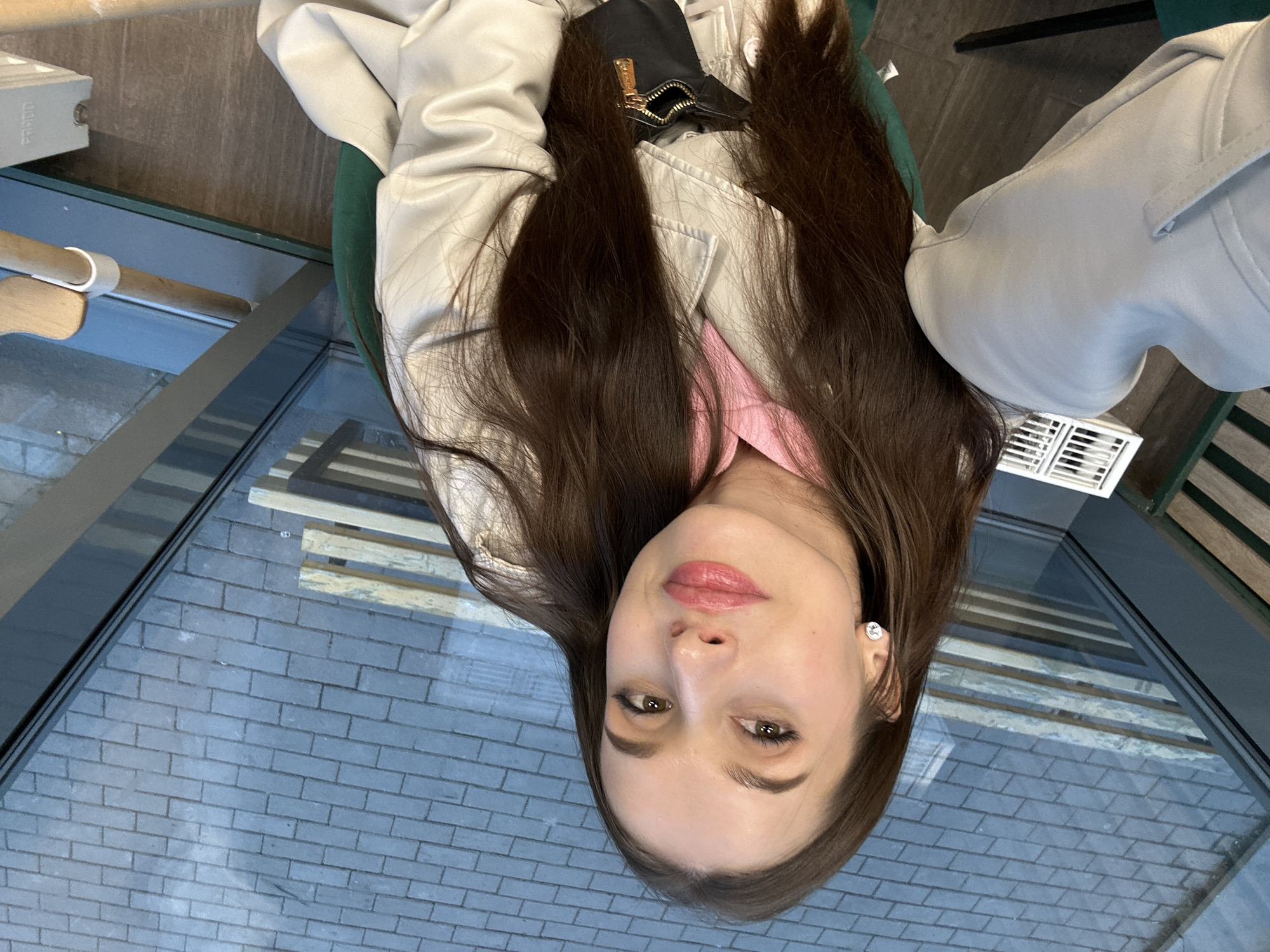 A woman with long, dark brown hair is posing in front of a large window with a view of a brick surface. The window reflects a pattern of parallel lines, potentially from a building or structure. The woman is wearing a light-colored jacket and a pink top underneath. Part of a black bag or purse is visible near her jacket. The location is possibly in Yekaterinburg, Russia, based on the surroundings.