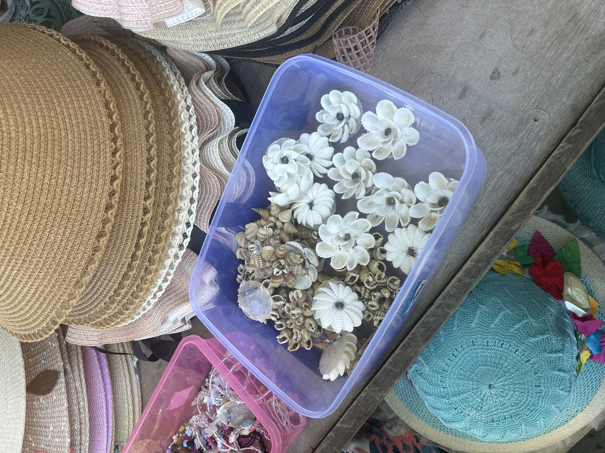 In Bira, Indonesia, on a weathered wooden surface, a stall displays various handmade crafts. Stacked straw hats in shades of beige and white dominate the upper portion of the image. In the foreground, a purple plastic container is filled with decorative items made from seashells. These include several flower-shaped ornaments and clusters of small shells arranged in circular patterns. To the left of the purple container, a pink plastic container holds a collection of colorful beads and small charms, possibly part of jewelry-making supplies. Below the containers, a light blue crocheted hat with a woven brim and decorative fabric flowers is visible. The overall scene suggests a market or craft stall offering beach-themed accessories.