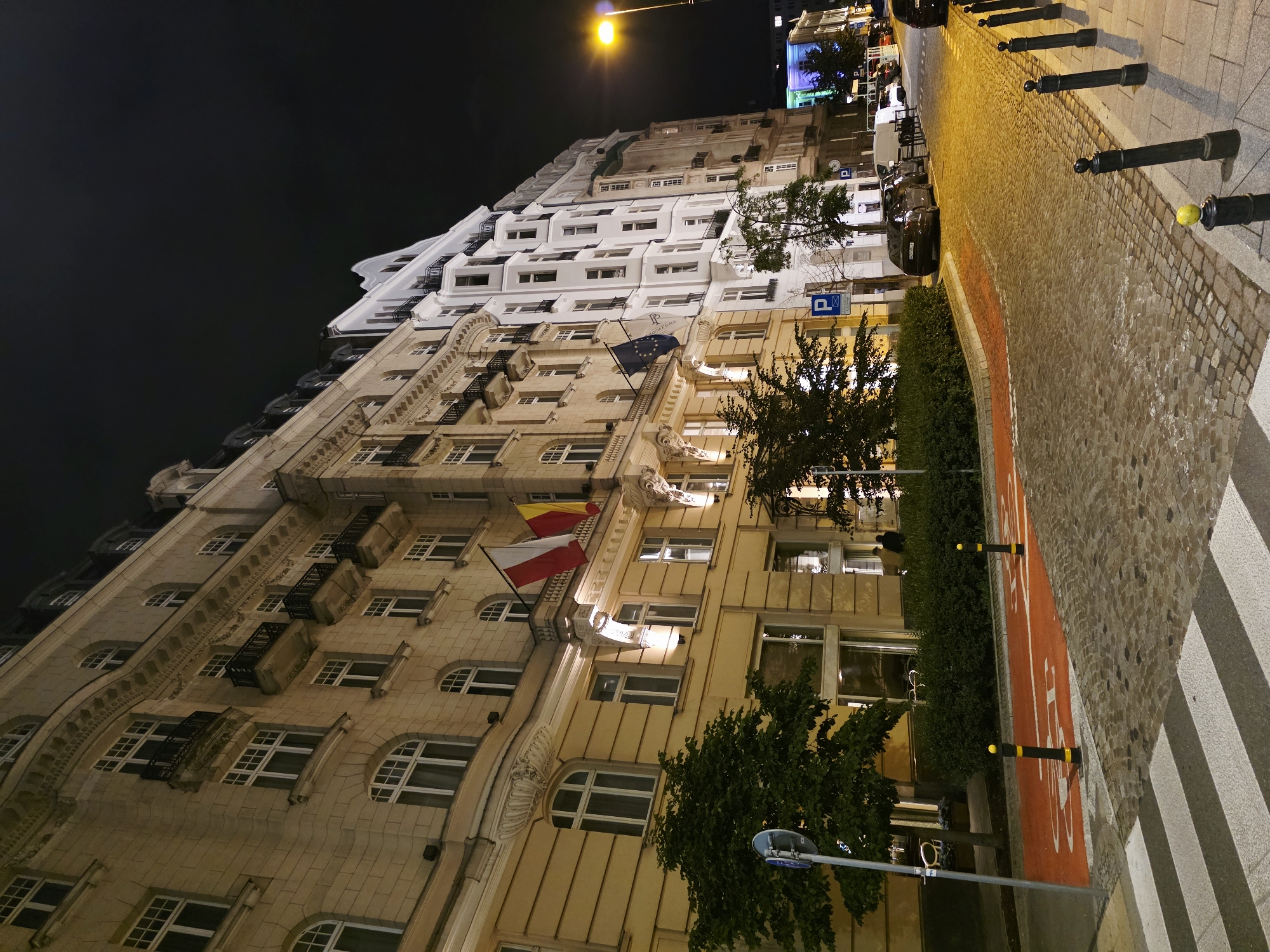 The image shows a street scene at night in Warsaw, Poland, featuring a row of historic buildings. The buildings are multi-story, with ornate architectural details, balconies, and arched windows. One building is a light beige color, while the adjacent one is white, creating a visual contrast. Flags are visible on the beige building, including what appears to be the Polish flag and another flag with yellow and red stripes.
The street is paved with cobblestones, and there is a designated bicycle lane marked with orange paint and white markings of a bicycle. A pedestrian crossing is also visible in the foreground. Several cars are parked along the street, and streetlights illuminate the scene with a warm, yellowish glow. Trees line the sidewalk and are interspersed with the buildings. The sky is dark, indicating nighttime. The overall impression is one of a well-preserved historic urban environment at night.