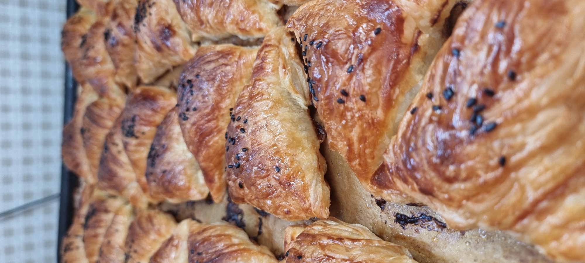 A close-up, eye-level shot displays a baking sheet filled with golden-brown, triangular pastries. The pastries, likely borek, are arranged in rows and appear to be fresh from the oven, with a glossy sheen and visible flaky layers. Black sesame seeds are sprinkled on top of many of the pastries. The background shows a blurred tiled surface, suggesting an indoor setting, possibly a kitchen or bakery. There is no discernible location in the city of Aktobe, Kazakhstan.