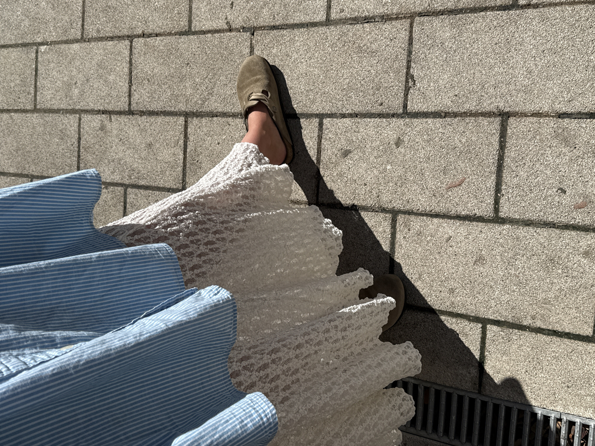 An eye-level, full-frame shot depicts a portion of a person standing on a paved surface, likely a sidewalk or pedestrian area. The person is wearing a blue and white striped shirt, a white, lacy skirt, and a light brown slipper or sandal on one foot. The paved surface consists of gray, rectangular bricks, laid out in a pattern. Strong sunlight casts shadows of the person and objects onto the pavement. A metal grate is visible at the edge of the frame. The location is suspected to be Belgrade, Serbia, although there is no explicit evidence in the image to confirm this.
