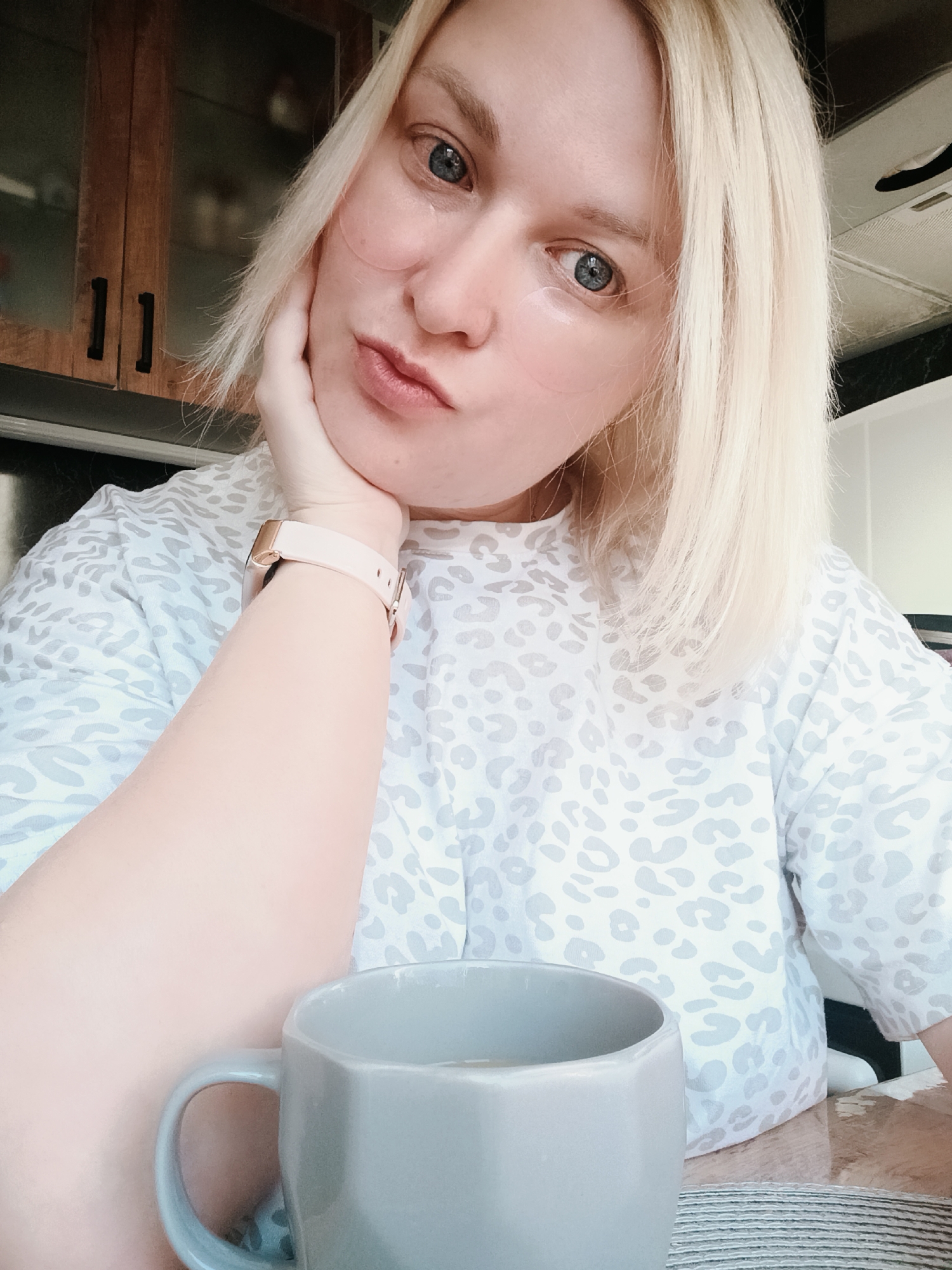 A person with light blonde hair and light skin poses for a selfie. They are wearing eye patches under their eyes and a light-colored shirt with a light gray leopard print. The person rests their face on their left hand, which has a pink smartwatch on the wrist. A gray geometric-shaped mug sits in the foreground. The background includes kitchen cabinets, a stainless steel refrigerator, and part of what appears to be a range hood. No specific features identify this as Chaikovsky, Russia.