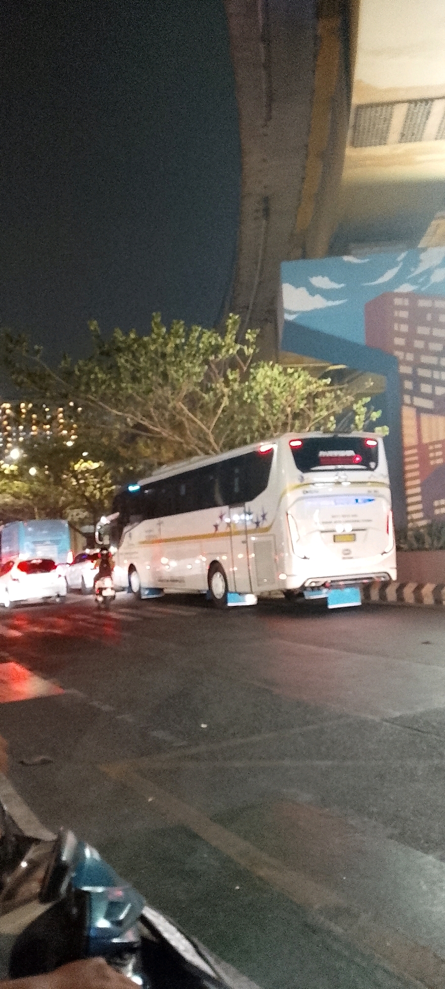 The image shows a street scene at night in the city of Bekasi, Indonesia. A white intercity bus is parked on the side of the road, with its lights on. The bus has yellow and blue accents and a logo with stars on its side. In the background, there is a mural depicting buildings and clouds on a wall. Trees with sparse foliage are visible behind the bus. Further down the road, two white cars are parked, and a scooter is moving towards the bus. The road surface is dark asphalt with visible lane markings. The lighting suggests it is nighttime, with artificial lights illuminating the scene.