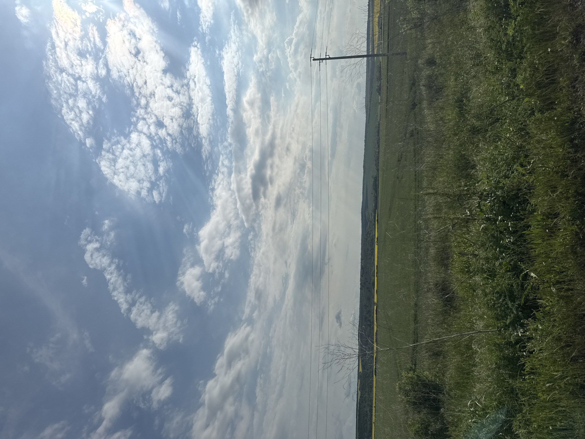The image shows a rural scene under a sky filled with scattered clouds and bright sunlight. The foreground consists of a field with varied green vegetation and some yellow flowering plants along its edge. An electrical power pole stands in the field, with power lines stretching across the view. The location is in Livada, Bulgaria. No people or animals are visible.