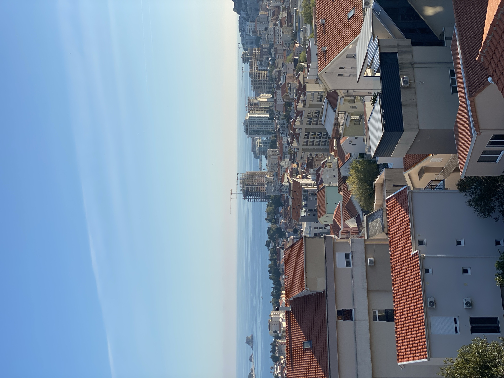 The image is a sideways aerial view of Budva, Montenegro. The Adriatic Sea stretches from the center towards the left. Residential and commercial buildings are densely packed with red-tiled roofs dominating the skyline. A construction crane is visible near a high-rise building under development. The sky is a clear, light blue.