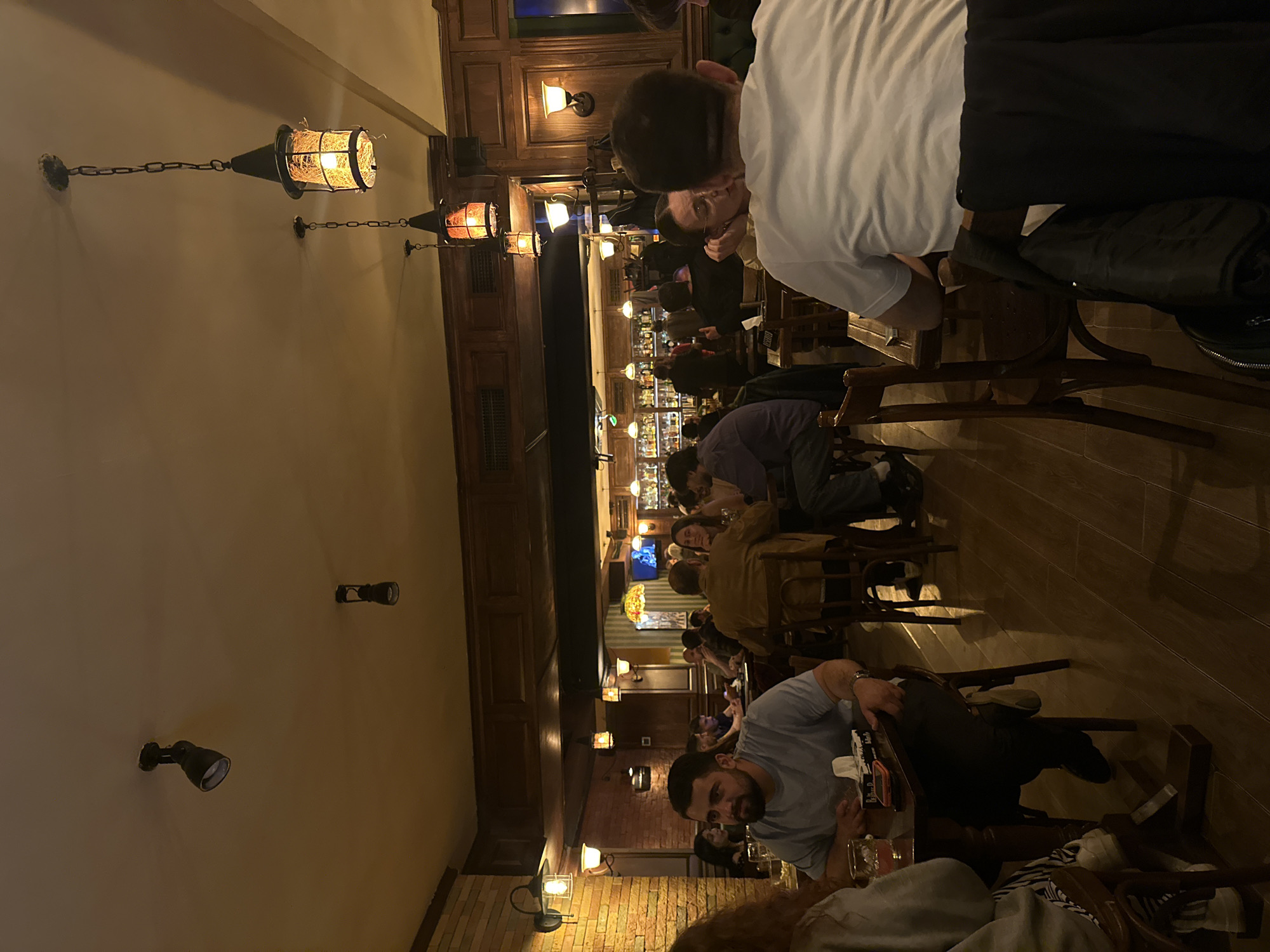 The photograph depicts the interior of a pub, seemingly located in Yerevan, Armenia, based on its aesthetic and probable contextual references. The scene is crowded, with patrons seated at tables stretching into the distance. Most of the people appear to be in casual attire, possibly enjoying a meal or drinks. The lighting is warm and somewhat dim, with decorative hanging lanterns casting light. There's a bar visible in the background with bottles and glassware. The furniture appears to be of dark wood, and the walls are paneled with dark wood and light colored exposed brick. Some people are engaged in conversations while others appear to be looking at their phones or menus.