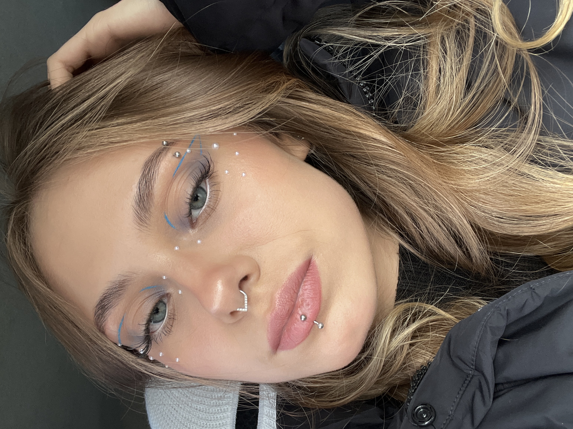 A close-up, angled portrait of a young woman with blonde hair, blue eyes, and fair skin. She is wearing makeup, including blue eyeliner and small pearl-like embellishments around her eyes and face. She has a nose piercing and a lip piercing. She is wearing a dark-colored puffy jacket. The location of the photo is unknown, and there is no evidence suggesting it was taken in Фоки, Russia.