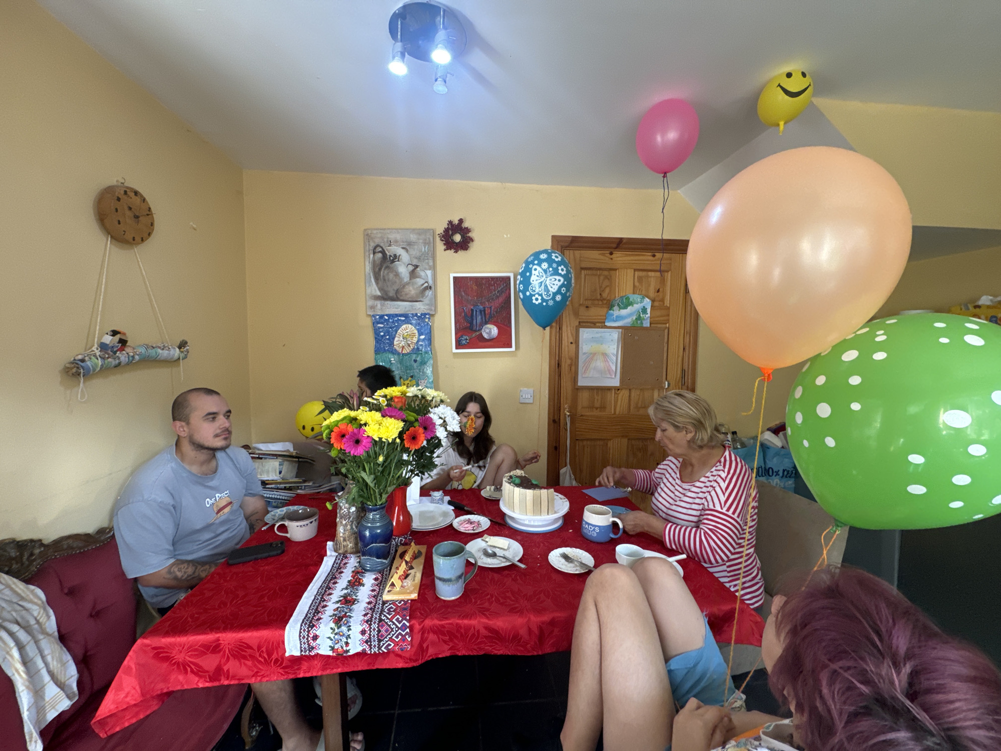 The image depicts an indoor scene of a family gathering, likely a birthday celebration, in a residential setting. The room appears to be a dining area, with a red tablecloth-covered table as the central focus. Several people are seated around the table, engaged in various activities.
On the left side of the frame, a man with a shaved head and tattoos is seated, wearing a grey t-shirt. A woman with blonde hair, wearing a red and white striped shirt, is seated opposite him, looking down at something on the table. A young woman with long brown hair is also at the table, and her legs are visible in the foreground, wearing blue shorts. Another person with dark, purple hair is partially visible in the lower right corner.
The table is laden with food and drinks, including a cake, plates with slices of cake, cups, and a vase of colorful flowers. A mobile phone is also visible on the table. In the background, there are several pictures on the wall, a clock, and balloons, including a yellow smiley face balloon, a pink balloon, a light blue balloon with a butterfly design, a large peach balloon, and a large green balloon with white polka dots. The walls are painted a pale yellow color, and the ceiling has modern lighting fixtures.
The overall impression is one of a casual and festive family occasion. The presence of balloons and a cake strongly suggests a birthday celebration.
This image is a still photograph of a domestic interior. There is no information in the image to suggest a specific location within the city of Waterford, Ireland.