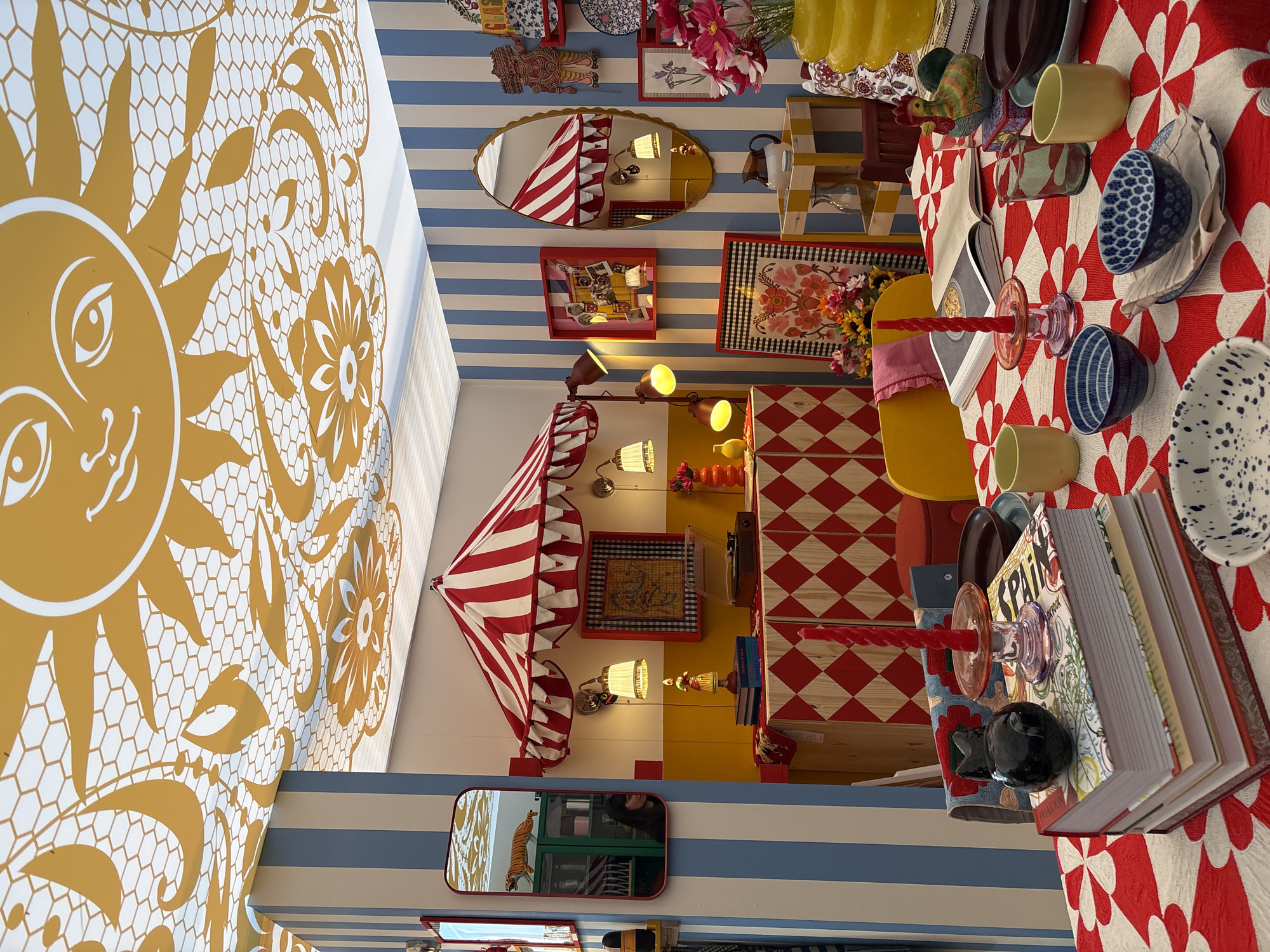 This vibrant image captures an elaborately decorated, whimsical interior space, likely an art installation or a themed room in Milan, Italy. The setting creates a surreal "upside-down" perspective, with furniture and decor arranged playfully as if defying gravity.
The room is characterized by a mix of bold patterns and colors. A striking blue and white horizontal striped pattern covers what appears to be both a wall and the floor. Other walls feature bright yellow sections and a red and white checkered pattern on a cabinet or wall panel. A prominent golden sun motif with a smiling face, set against a white background with intricate lace-like floral and honeycomb patterns, dominates a large surface, suggesting it could be a ceiling or large wall mural.
An "inverted" dining table, adorned with a red and white radial patterned tablecloth, holds various ceramic dishes including blue and white patterned bowls, speckled white bowls, and yellow cups. A clear glass and a striking red twisted candle in a glass holder are also present. A large, open cookbook, with "SPAIN" and "PHAIDON" visible on its spine, rests on the table. A yellow chair is positioned alongside the table.
Other notable objects include several mirrors, one oval mirror reflecting a red and white striped tent-like awning, and another rectangular mirror showing a tiger statue and a partial reflection of a person. Framed artworks, including one with a black and white checkered border and floral design, and another featuring old photographs, adorn the walls. A ceramic chicken figurine, potted flowers, a record player, and various lamps with warm yellow light further populate the space, contributing to a maximalist and lively atmosphere. No specific activity is taking place; it is a static, highly stylized display evoking a sense of playful surrealism.