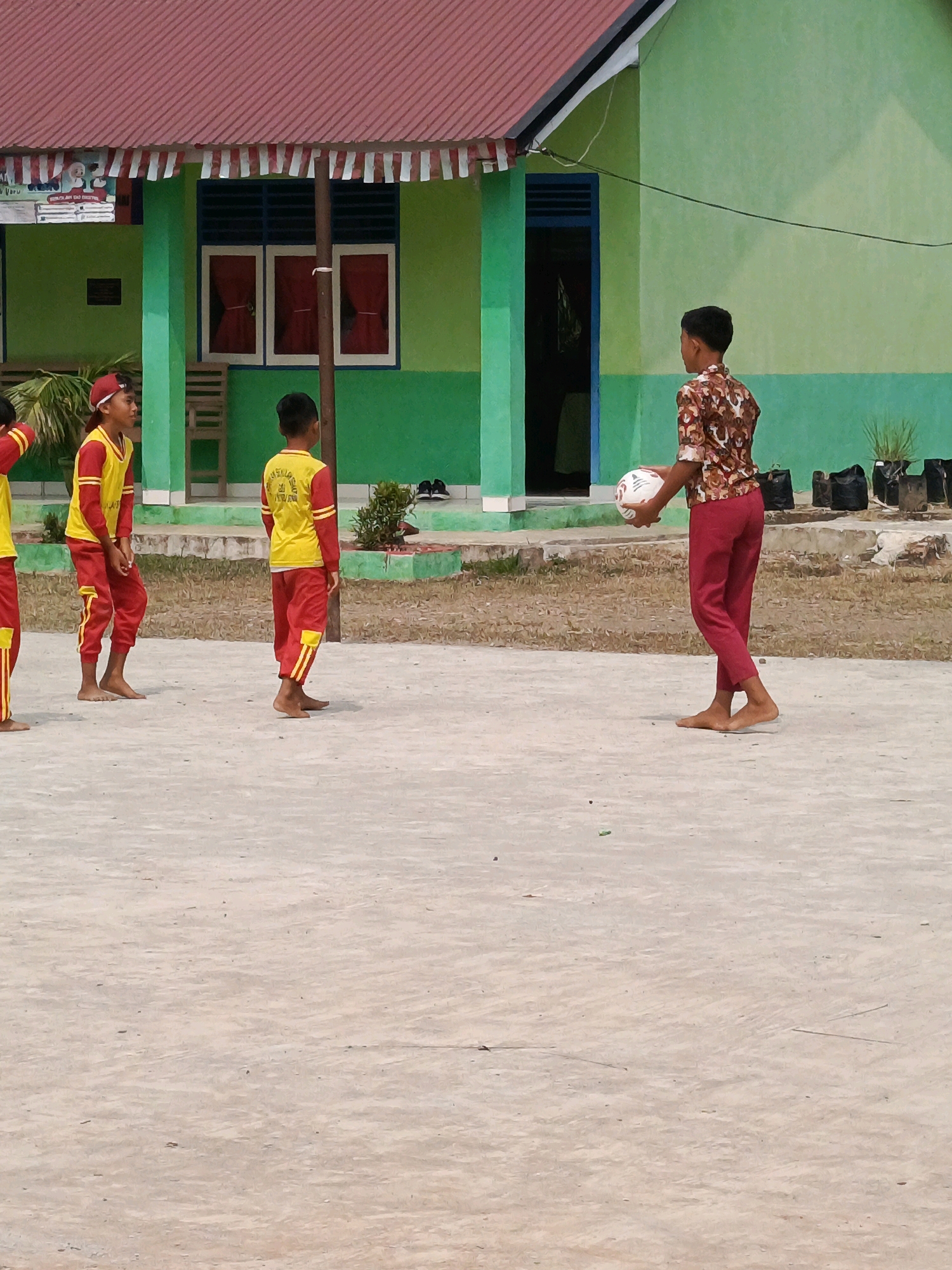In Megang Sakti IV, Indonesia, a group of children are engaged in an outdoor activity on a paved court in front of a school building. The school's exterior features a green painted facade with a red-tiled roof. Three children, all wearing red pants and yellow shirts, are visible. One child, on the far left, is partially visible and appears to be wearing a red cap. The middle child, also in red and yellow attire, is standing barefoot, looking towards the right. The child on the right, also in red pants and a yellow shirt, is holding a white and red soccer ball with both hands, appearing ready to play. Another child, wearing a patterned brown shirt and red pants, is also barefoot and standing to the right of the other children, holding the soccer ball, implying an interaction or game is in progress. The environment appears to be dry with scattered patches of dry grass and some black potted plants in the background. The ground is a concrete surface.