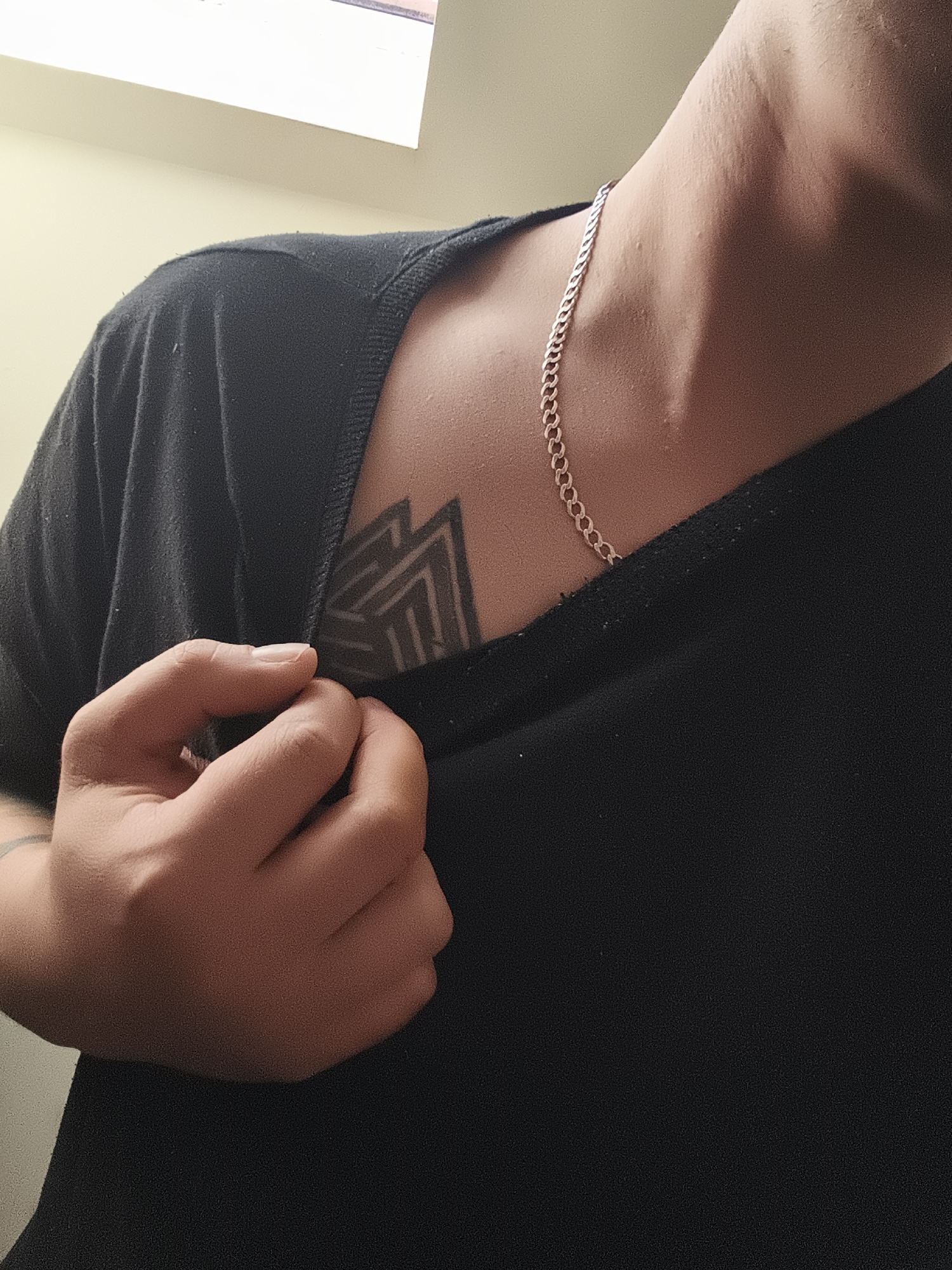 A close-up shot focuses on the upper torso of a person wearing a black t-shirt and a silver chain necklace. A tattoo, composed of geometric shapes, is visible on the upper chest, partially obscured by the shirt's neckline. The person's left hand is slightly raised, holding a section of the t-shirt. The background includes a portion of a light-colored wall and a window, suggesting an indoor setting. The location in the city of Nowy Tomyśl, Poland is not discernible from this image.