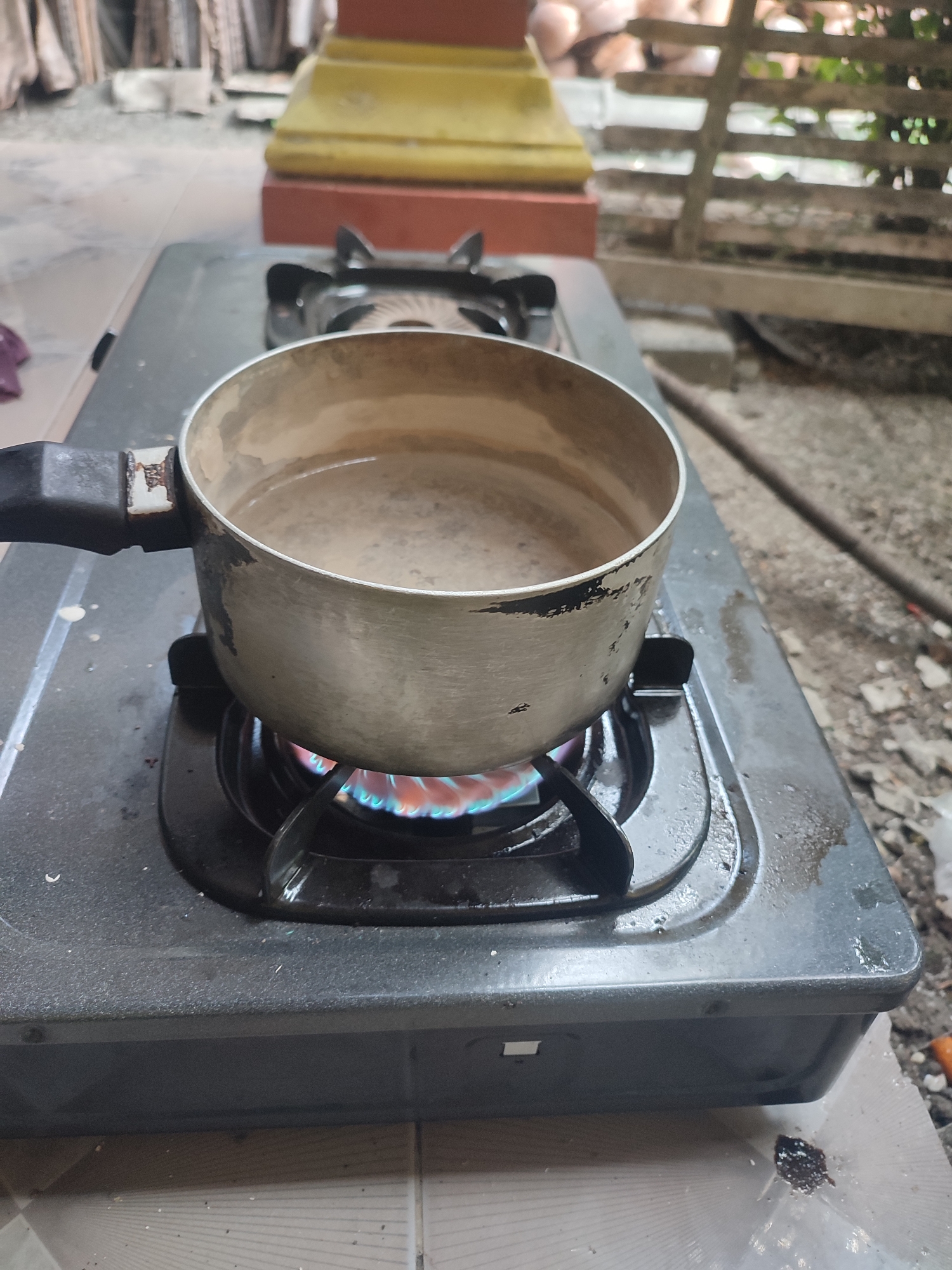 A close-up, eye-level shot shows a pot of light brown liquid being heated on a black gas stove. The pot is made of metal and appears slightly worn, with some black markings around the rim. The flame from the stove is visible underneath the pot, showing blue and orange hues. The stove itself is placed on a tiled surface. In the background, out of focus, are wooden slats, possibly part of a fence or a building structure, and some blurry yellow and red objects. The overall setting suggests an outdoor or semi-outdoor environment, common in residential areas of Sarwadadi, Indonesia.