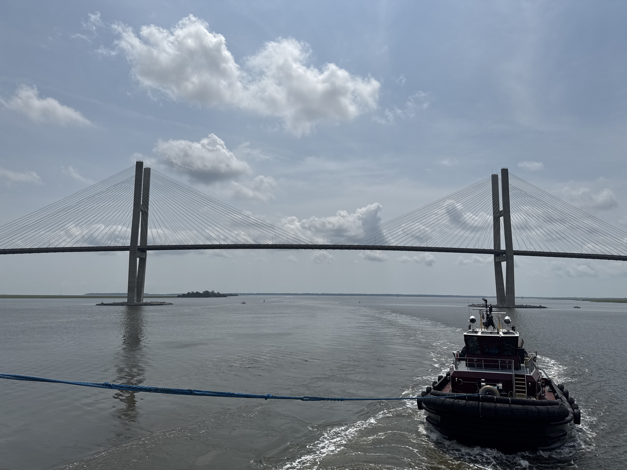 The image depicts a scene on the Brunswick River in Brunswick, Georgia, United States of America, under a partly cloudy sky. In the foreground, a red and black tugboat is moving away from the viewer, creating a white wake in the choppy grey water. A thick blue rope connects the tugboat to the camera's perspective, suggesting the camera is on another vessel.
Dominating the midground and background is the massive Sidney Lanier Bridge, a cable-stayed bridge with two tall, concrete towers. The bridge spans the river, with its roadway and numerous cables visible. The structure casts a reflection on the water below. On either side of the river, flat, tree-lined shores are visible. In the distance, on the horizon, a few smaller boats can be seen on the water, and the sky is filled with scattered white clouds against a pale blue backdrop. The overall impression is one of maritime activity under a typical summer day's sky.