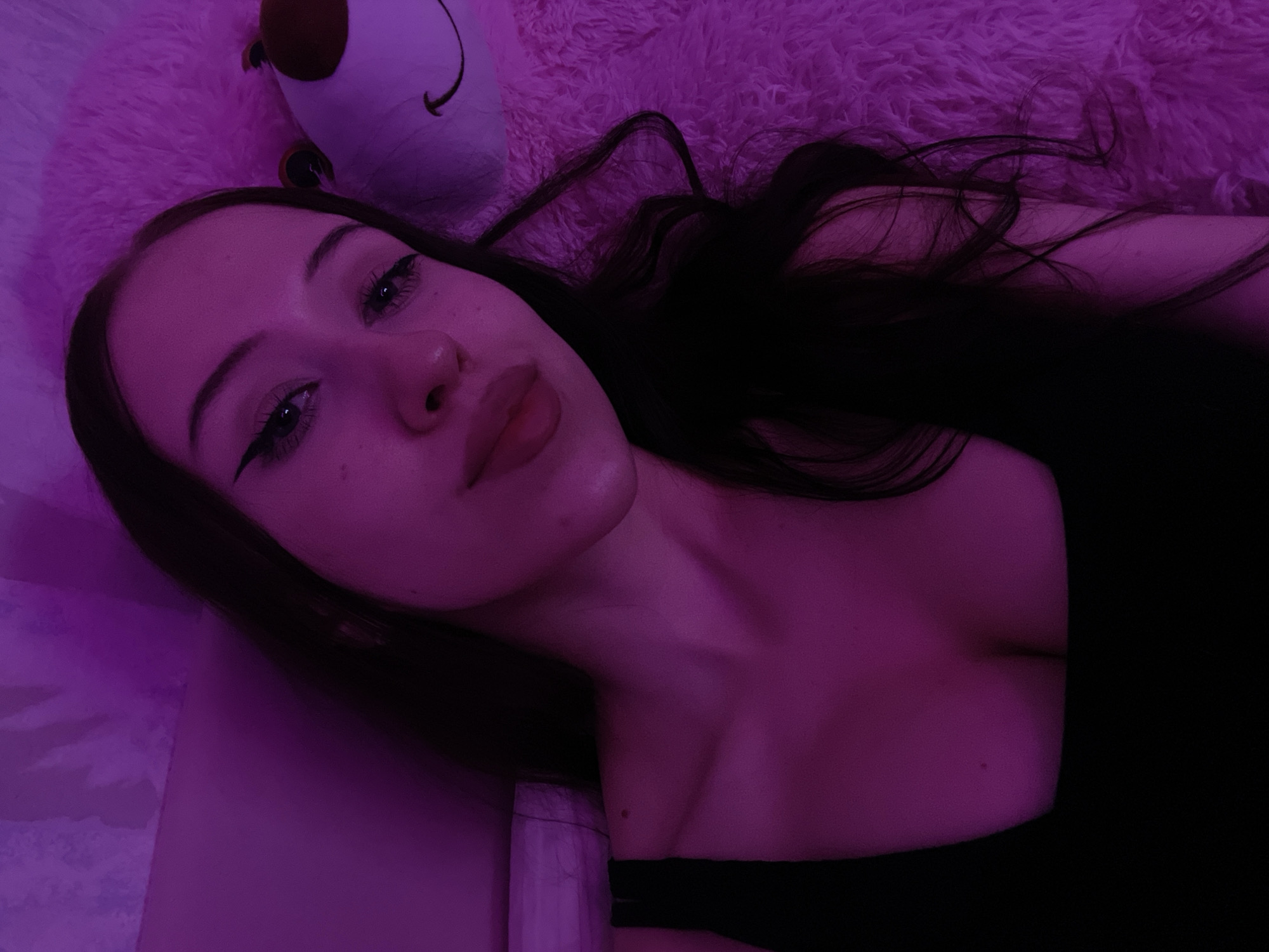 A young woman with long, dark hair, wearing a black tank top, is lying down. She has dramatic winged eyeliner and full lips. She is positioned next to a large, fluffy pink stuffed animal, possibly a bear, which has a smiling face visible. The scene is illuminated by purple lighting, creating a moody ambiance. The background appears to be a pink, textured surface, possibly a blanket or rug. The image is a close-up selfie, focusing on the woman's face and upper body. The location is not specified in the image.