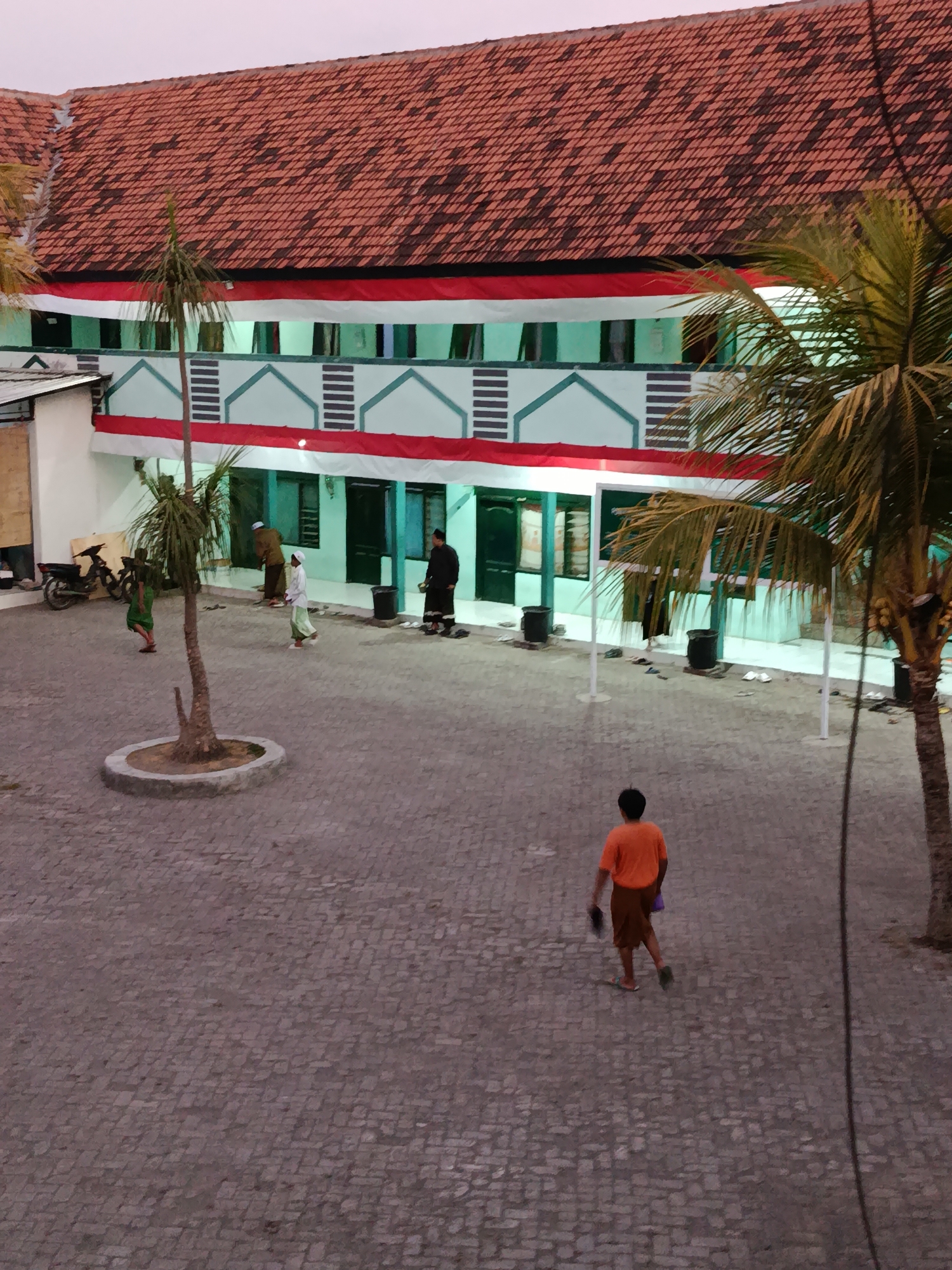 This is an eye-level, medium shot of a courtyard in front of a building in Ramban Wetan, Indonesia. The building has a red tile roof and is decorated with red and white banners along the second story. The building appears to be a school or religious institution, with multiple doorways and windows visible. In the foreground, a young person wearing an orange t-shirt and brown shorts walks away from the camera, holding a dark object in their right hand and a purple bag in their left. Several other individuals are visible in the mid-ground, some appearing to be praying or walking. A motorcycle is parked to the left of the building. The courtyard is paved with cobblestones. Two palm trees are present in the scene, one in the mid-ground and another to the right of the frame. The sky is overcast and light.