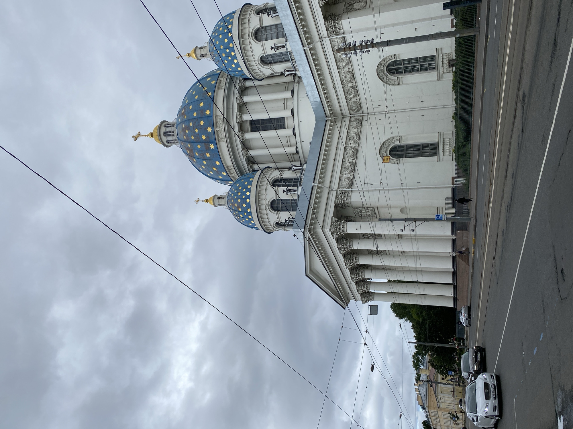 The image shows a view of the Trinity Cathedral in Saint Petersburg, Russia. The cathedral, a prominent landmark, features large white walls and multiple blue domes adorned with golden stars. In the foreground, a wide road with tram tracks runs horizontally across the frame. Several cars are visible on the road, including a white Jaguar in the left lane, a dark sedan, and another white car further down. A person is walking on the sidewalk on the right side of the road, and there are parked cars and streetlights visible. Overhead, a network of electrical wires crisscrosses the cloudy sky, partially obscuring the view of the cathedral's upper sections. The architecture is neoclassical, with a colonnade of tall white columns supporting a pediment. The sky is overcast with a grey, cloudy atmosphere.