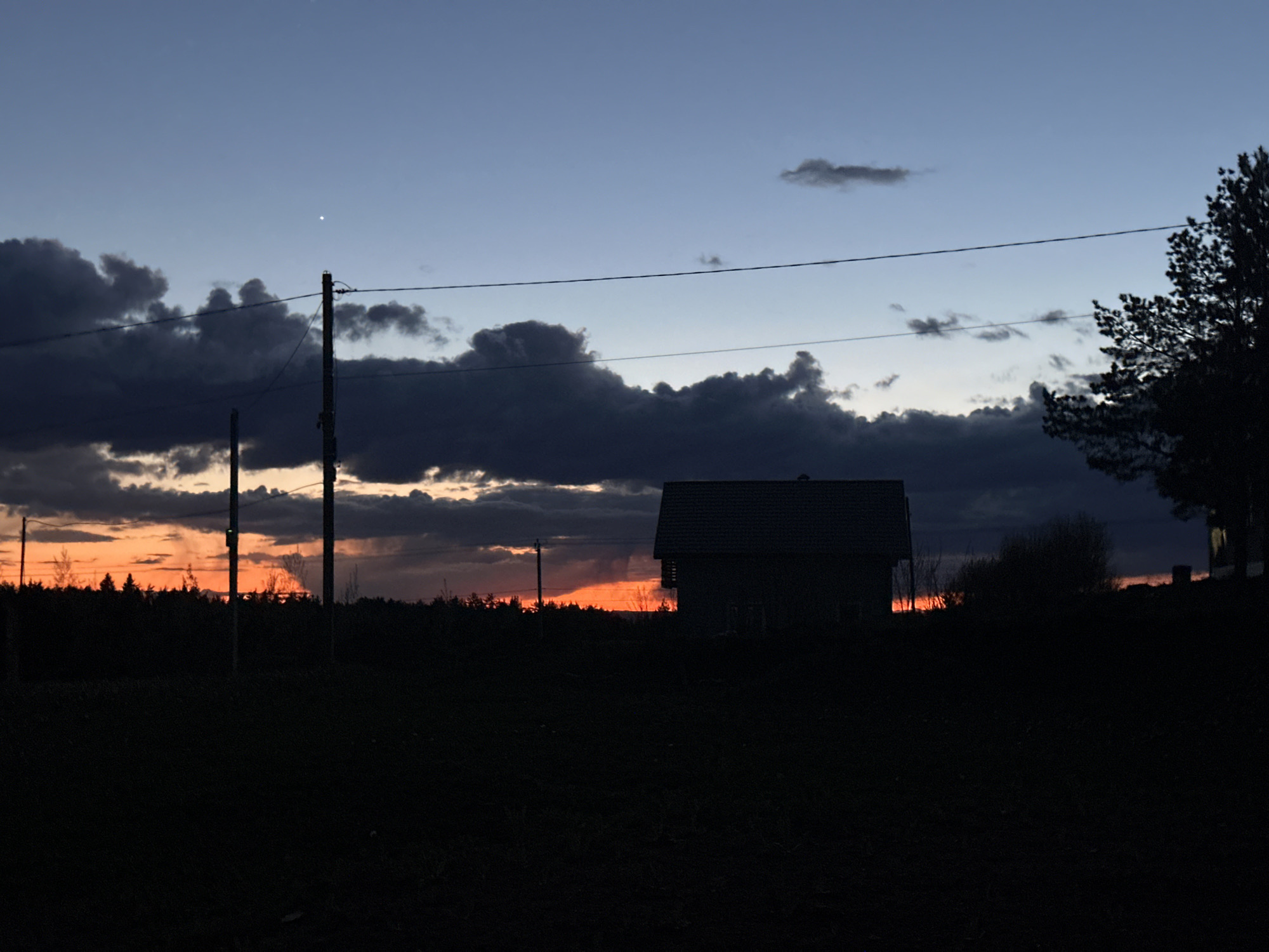 The image depicts a twilight or pre-dawn scene in Прикамский, Russia, evidenced by the presence of a building and power lines, elements commonly found in populated areas. The foreground is predominantly dark, suggesting low lighting conditions. A building, likely a house, is silhouetted against the horizon. The sky transitions from a dark blue at the top to a vibrant orange near the horizon, indicative of a sunrise or sunset. Dark clouds are scattered across the sky, obscuring parts of the colorful horizon. A power line stretches across the frame, punctuated by two utility poles. Silhouetted trees or bushes appear in the mid-ground.