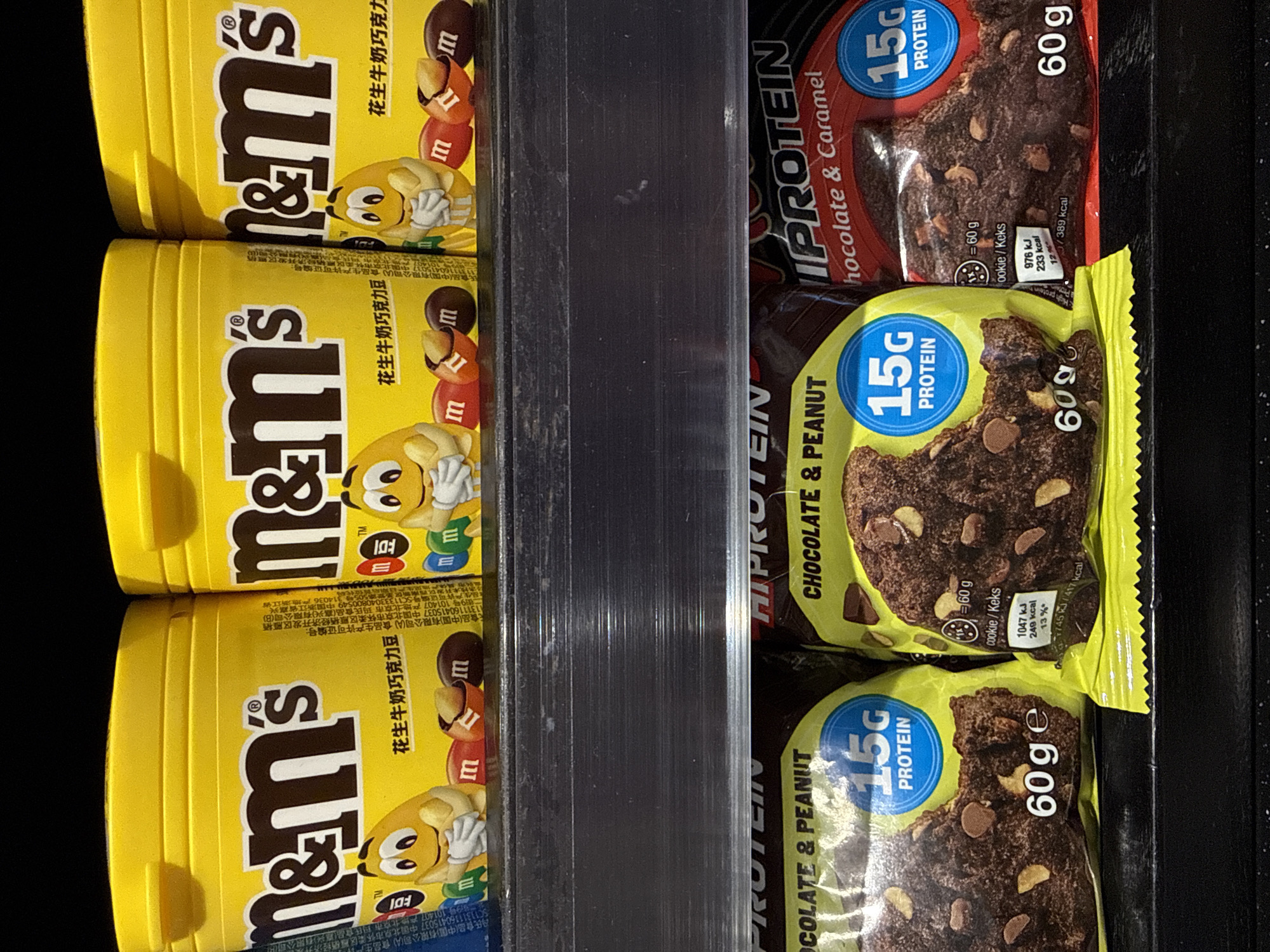 The image shows a shelf stocked with two types of snacks: M&M's peanut-flavored chocolate candies in yellow cylindrical containers and HiProtein chocolate bars. The M&M's containers are stacked vertically. The HiProtein bars are packaged in wrappers with blue and red accents, advertising "15G PROTEIN" and a weight of "60 g". The flavors shown are "CHOCOLATE & PEANUT" and "Chocolate & Caramel". The background is dark and out of focus. The location appears to be a retail environment, possibly a convenience store or supermarket.